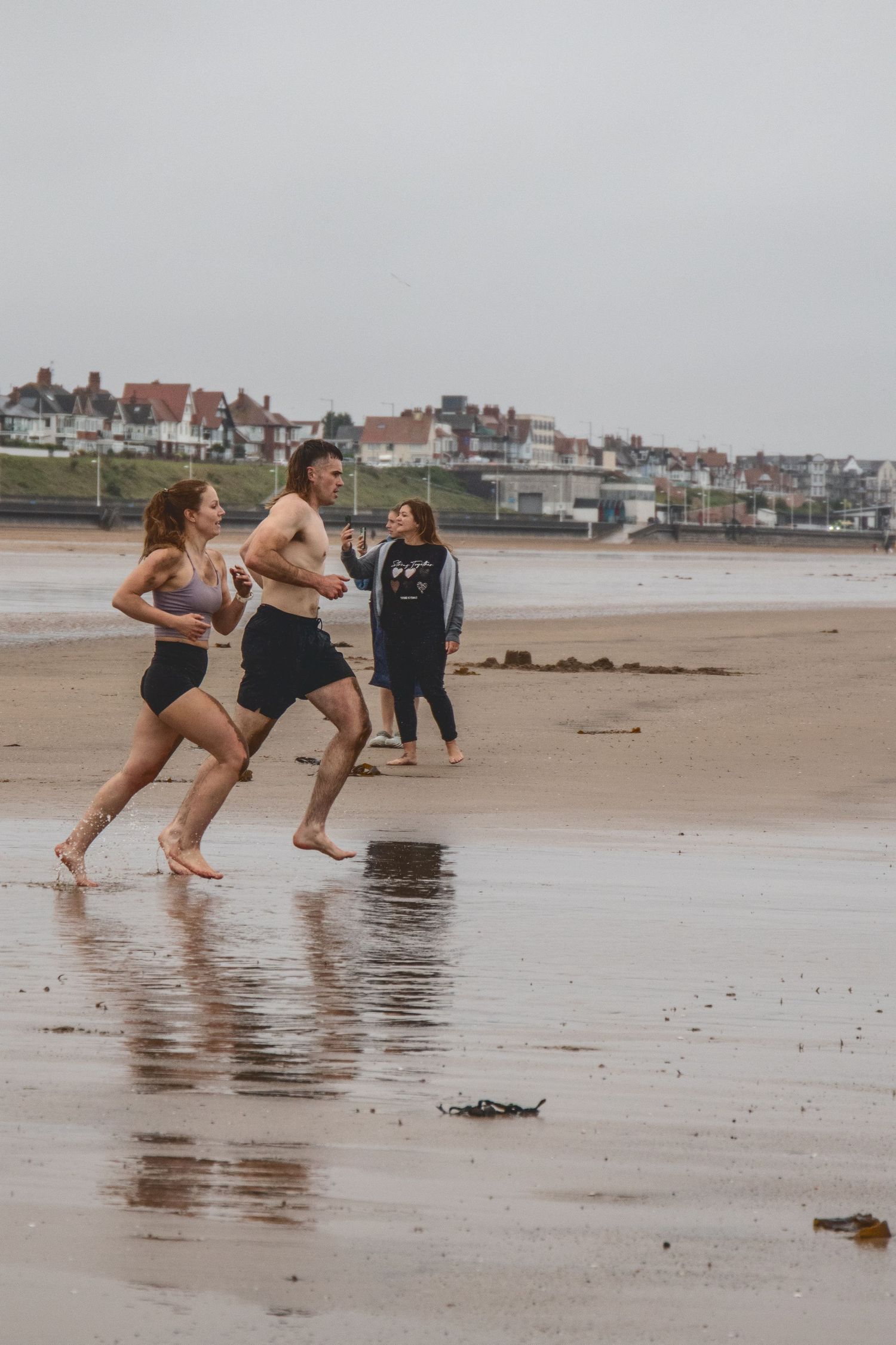 People jog along a misty beach with residential buildings visible on the coastline in the background.