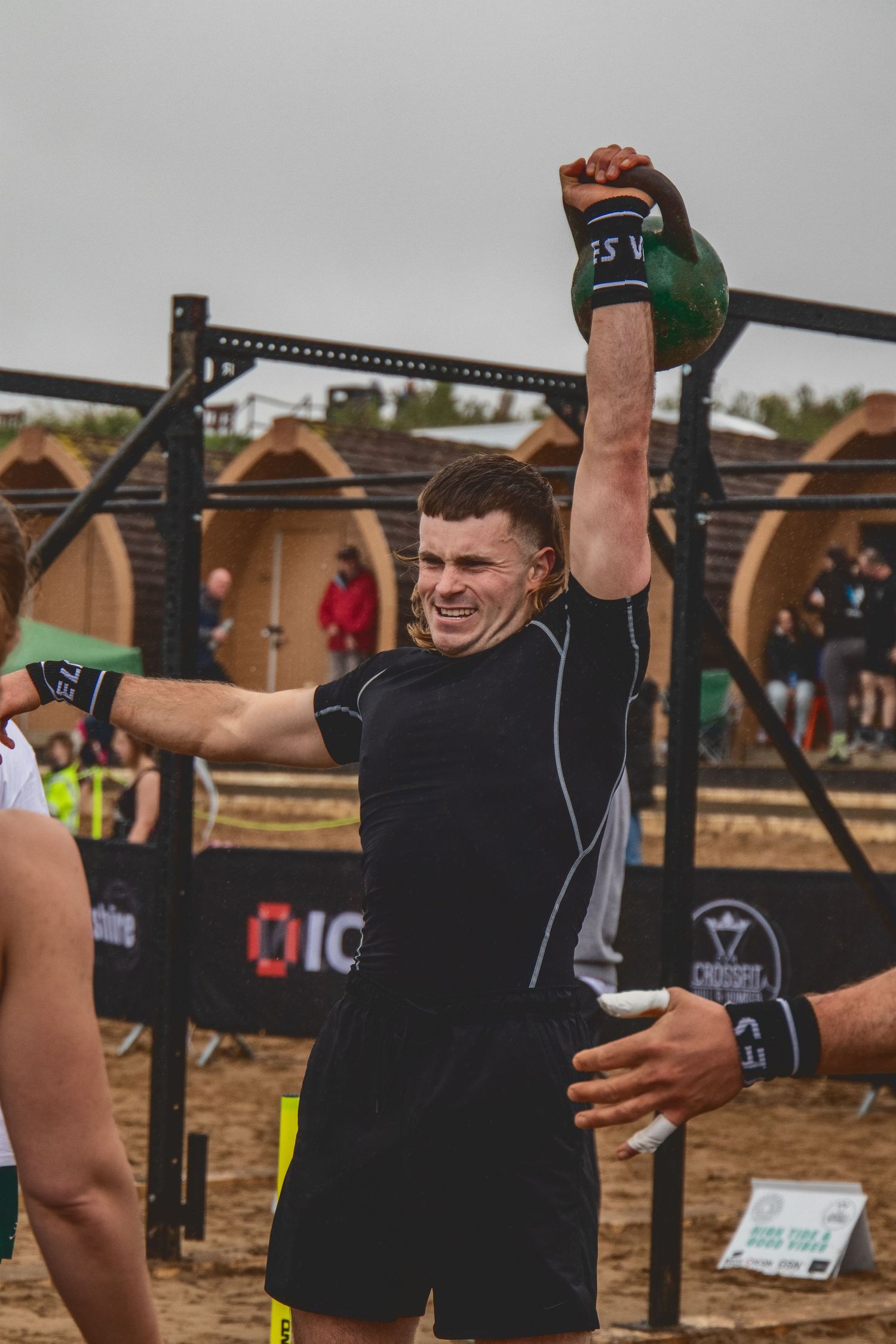 Athlete wearing black compression gear performs a kettlebell overhead press during an outdoor competition event.