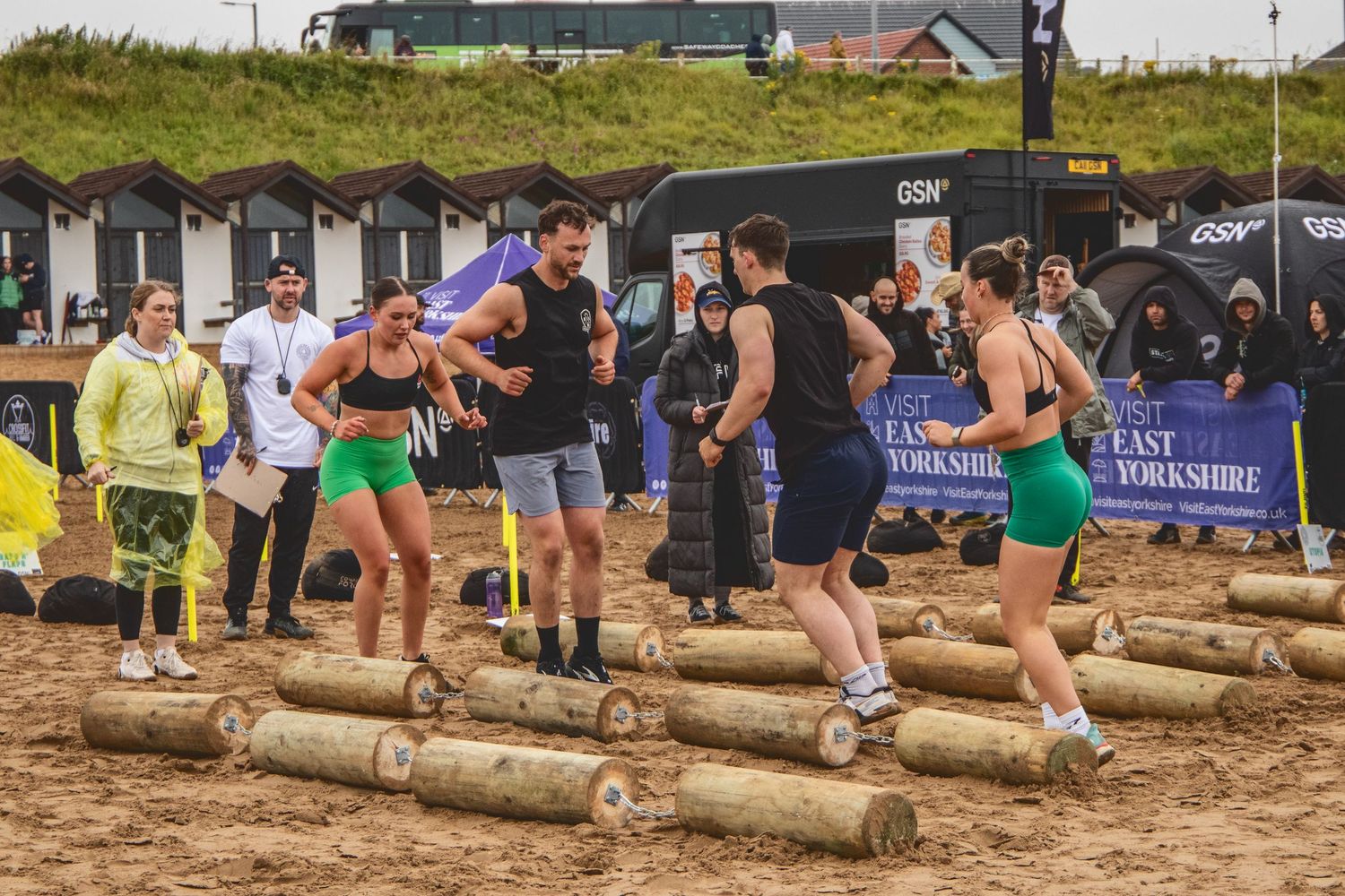 Competitors race across lined up logs during an outdoor obstacle course competition on sandy terrain.