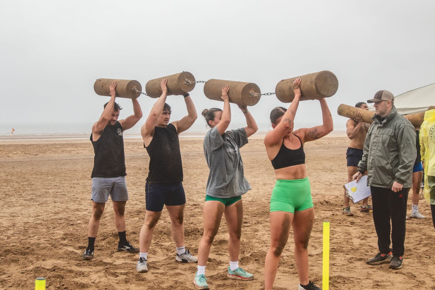 Group exercising with heavy wooden logs during an outdoor training session on a misty beach.