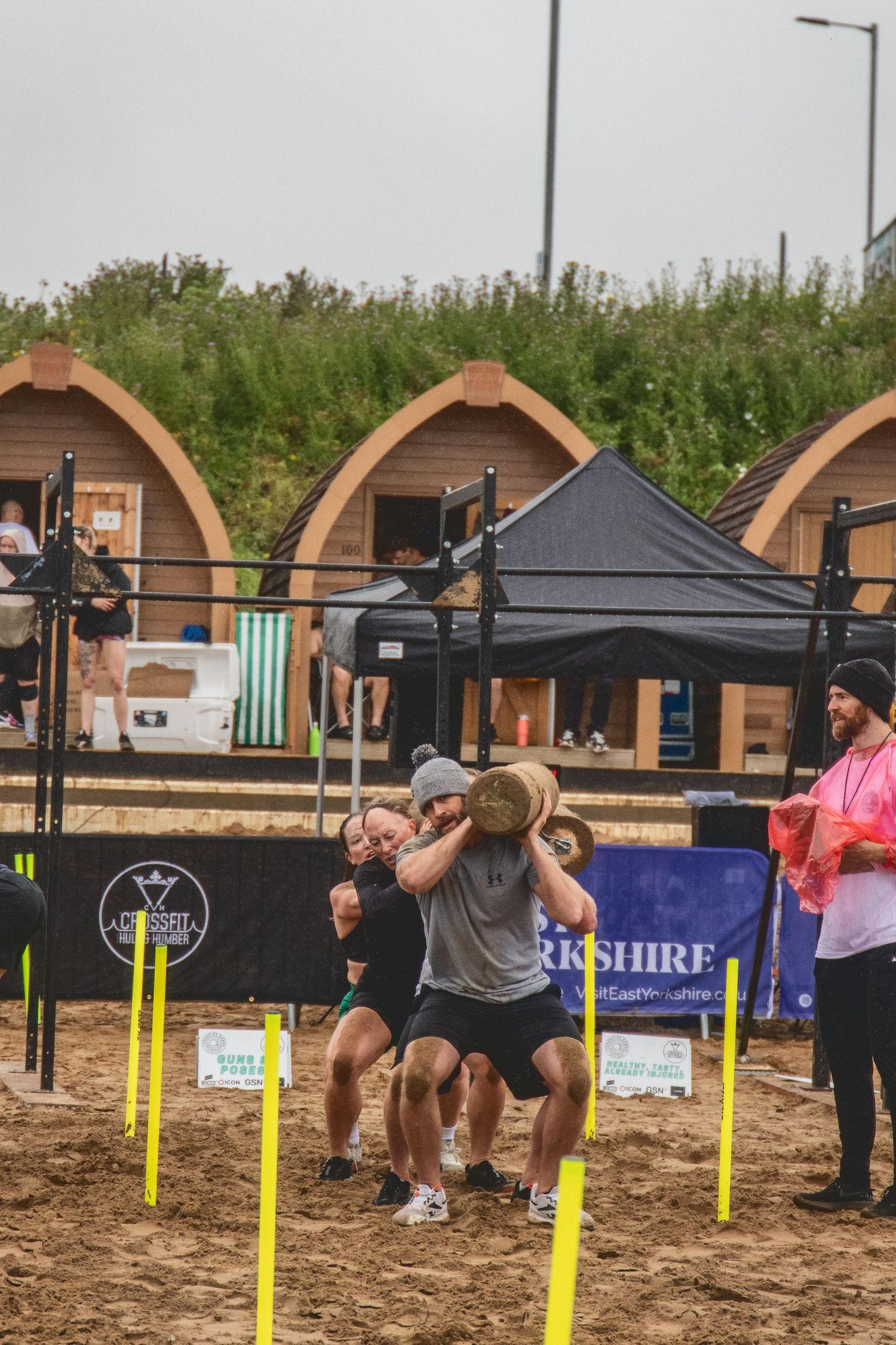 Athletes compete in a fitness competition with sandbags near wooden arch-shaped cabins and competition equipment setup.