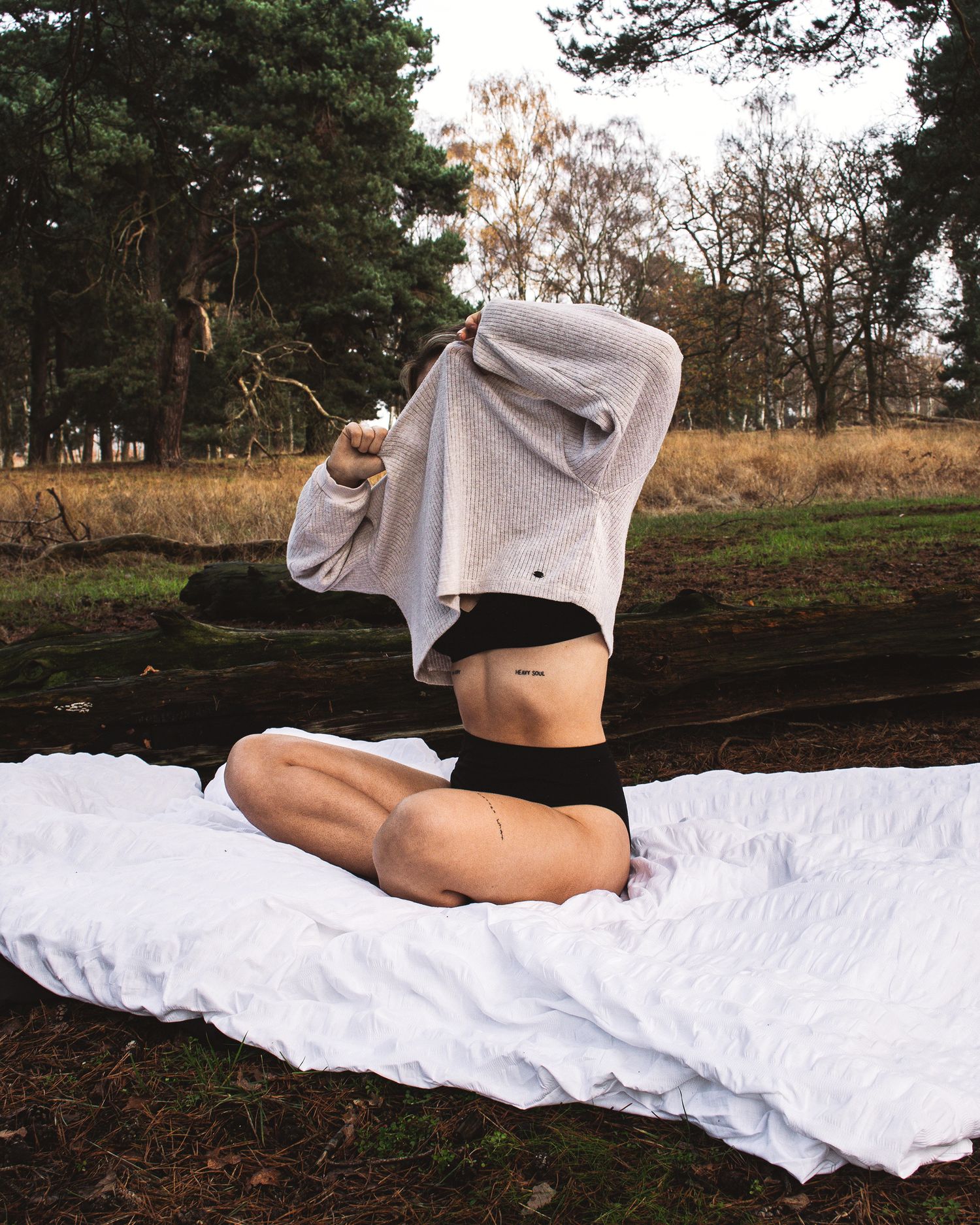 Person in light clothing lying on white blanket outdoors with pine trees in background.