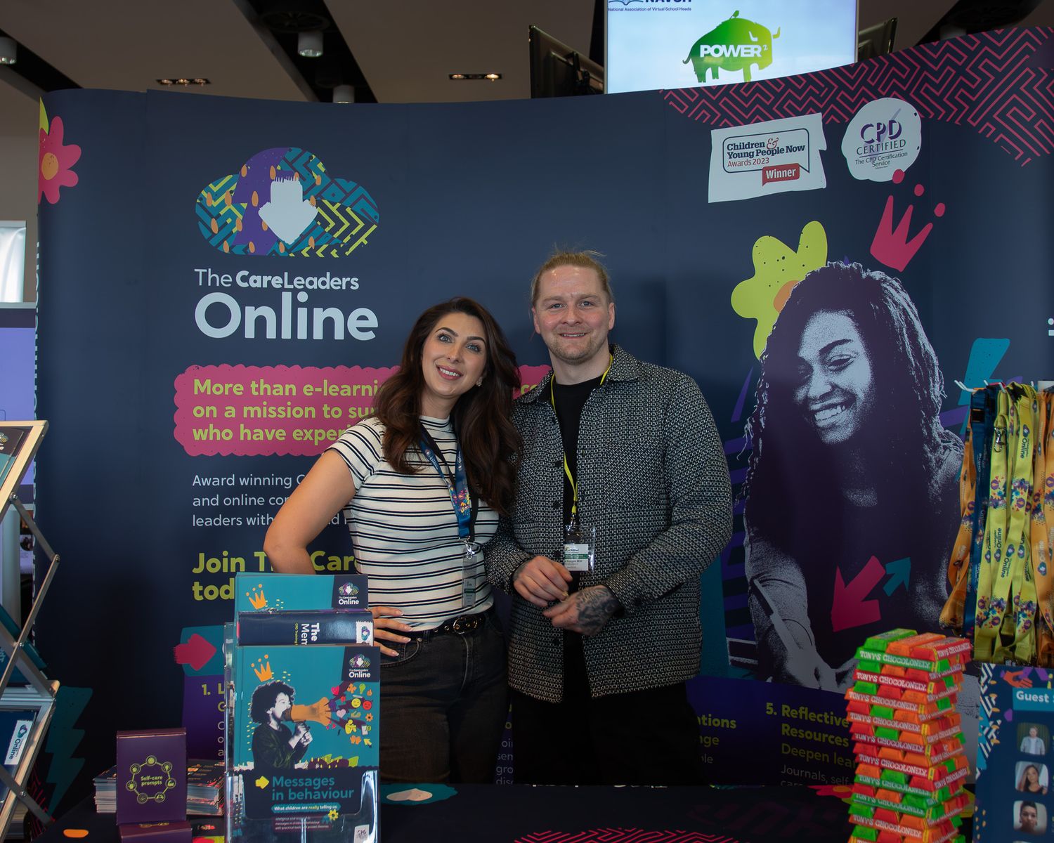 Two individuals stand at a promotional booth with colorful displays and stacked products at a marketing event.