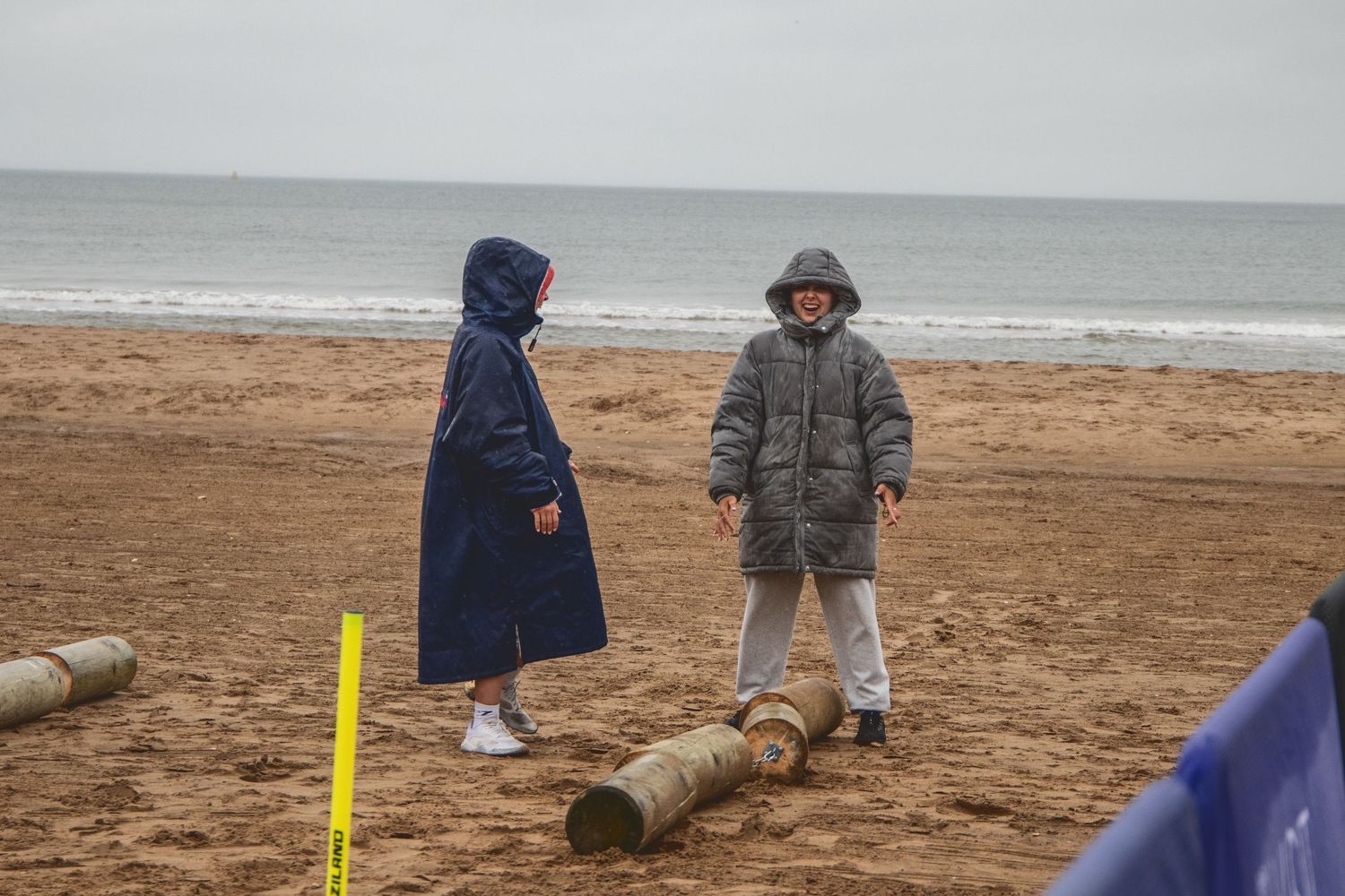 People in winter coats stand on logs at an overcast beach with yellow markers in the sand.