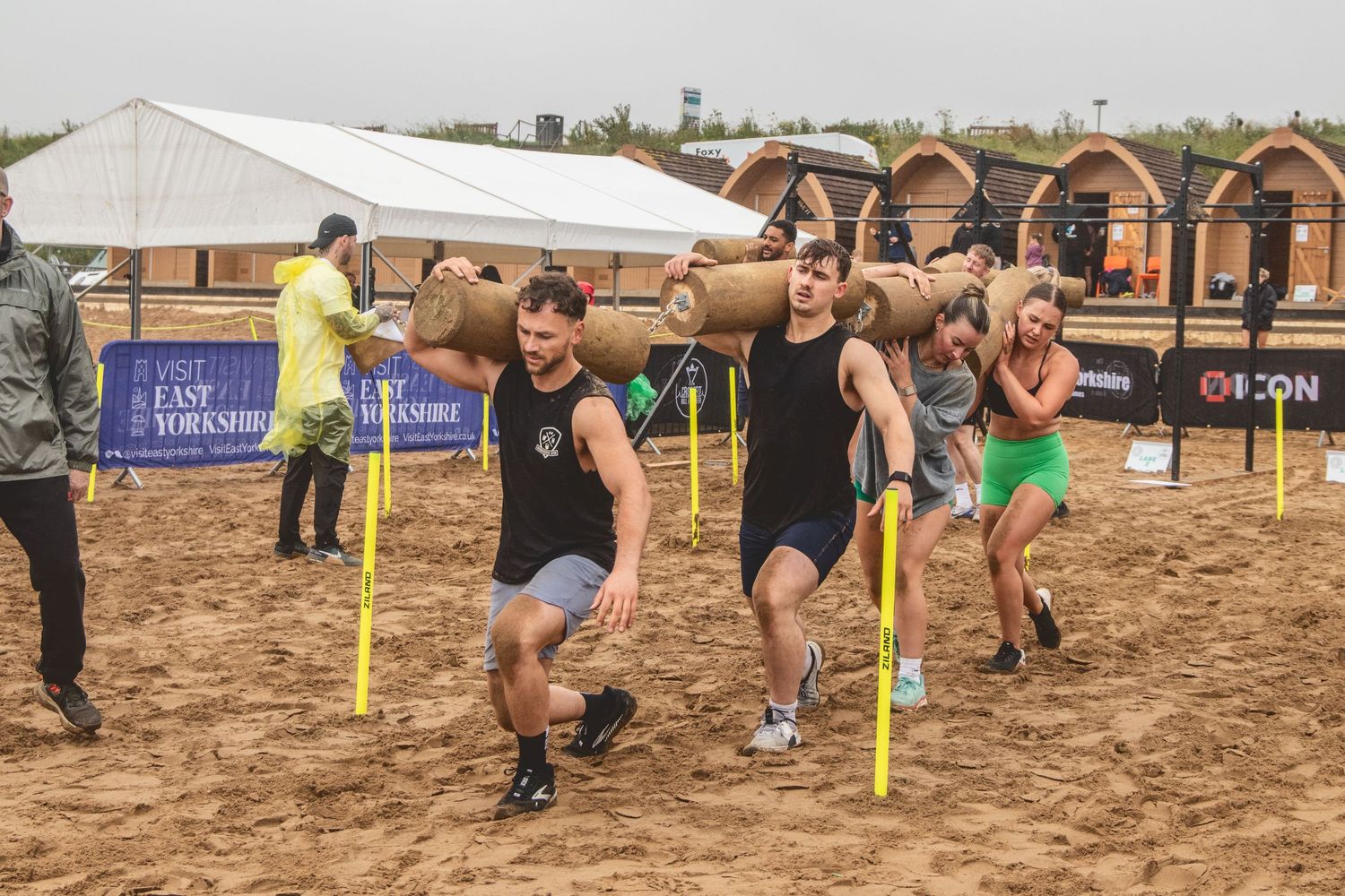 Athletes competing in a beach obstacle course race sprint together through poles on sandy terrain under a white canopy.