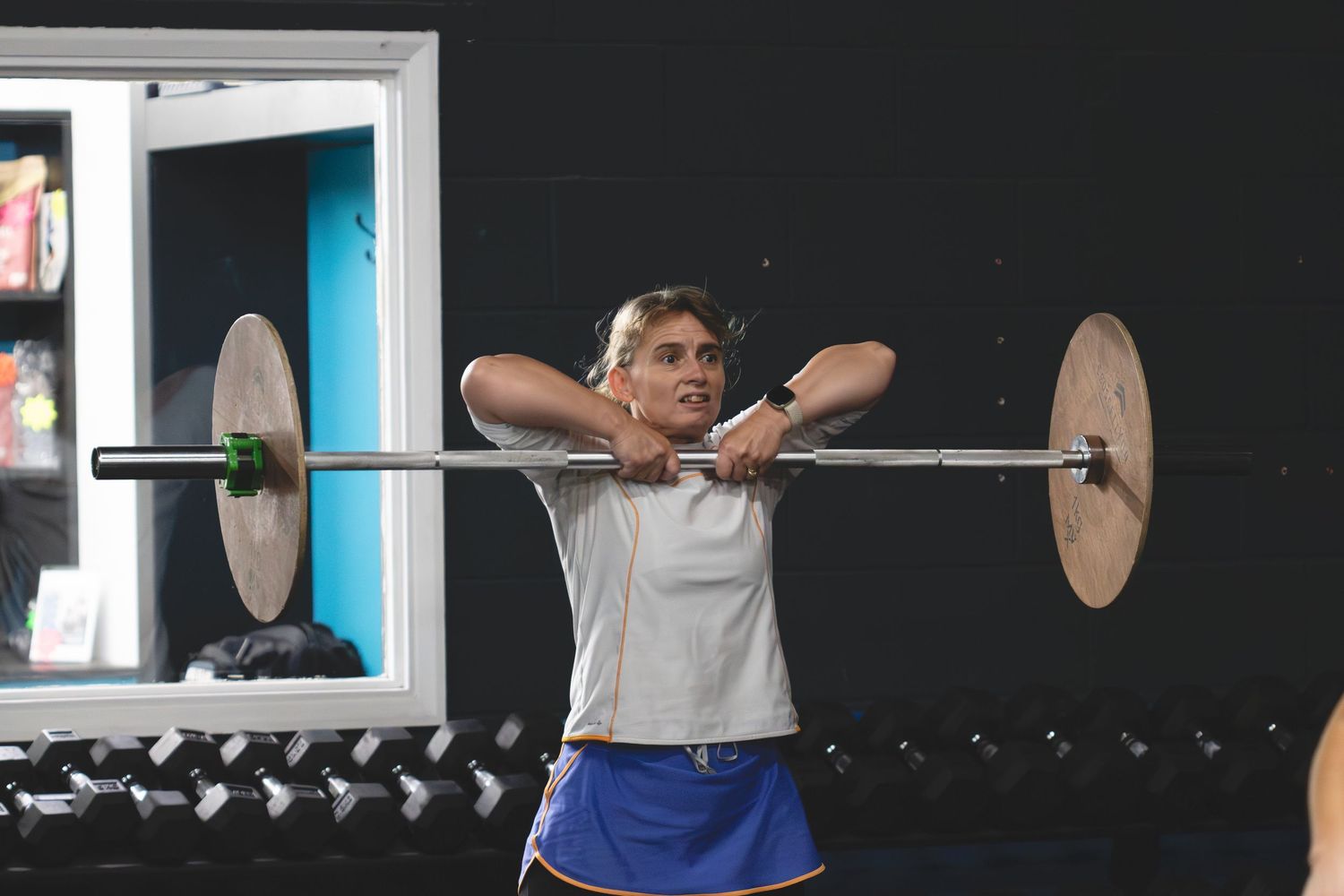 A person performing an overhead barbell exercise in a gym with dumbbells visible in the background.