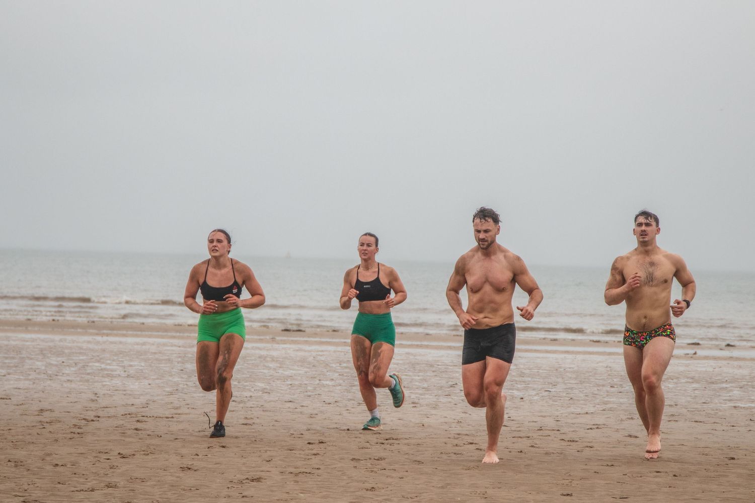 Four shirtless people running on a foggy beach wearing shorts and swimwear during exercise training.