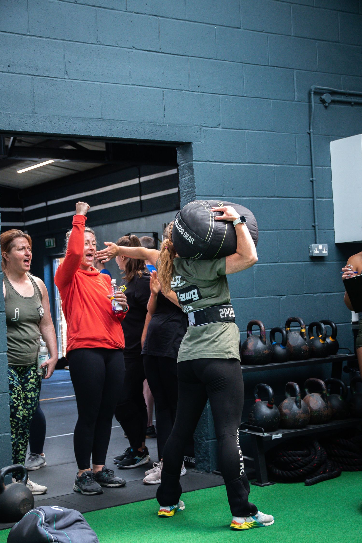 Fitness class participants workout with kettlebells in a gym with green flooring and gray walls.