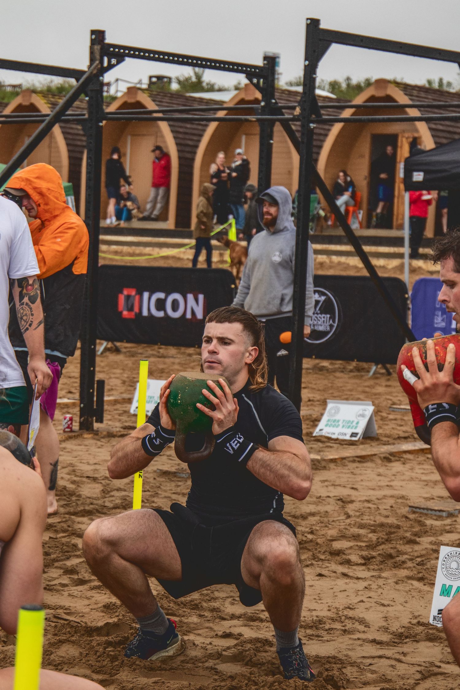 Athletes perform squats with medicine balls during an outdoor fitness competition on sandy terrain near workout equipment.