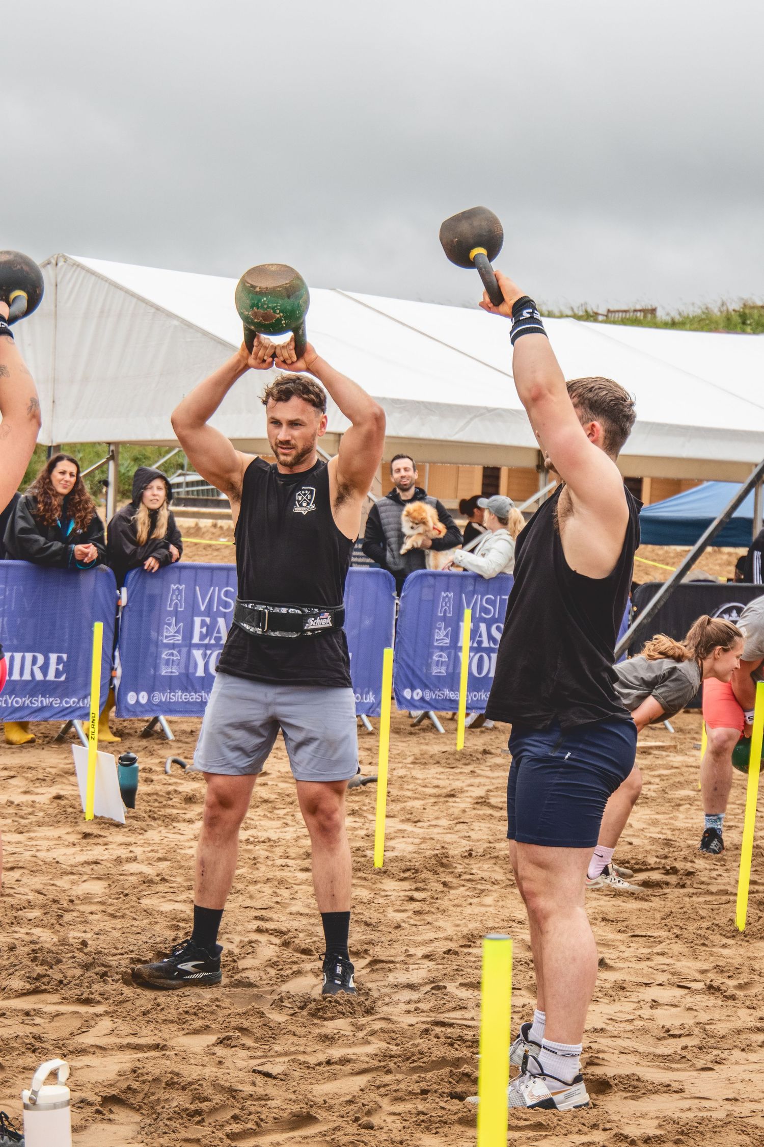 Athletes lifting kettlebells during an outdoor competition on sandy terrain with spectators and banners visible.