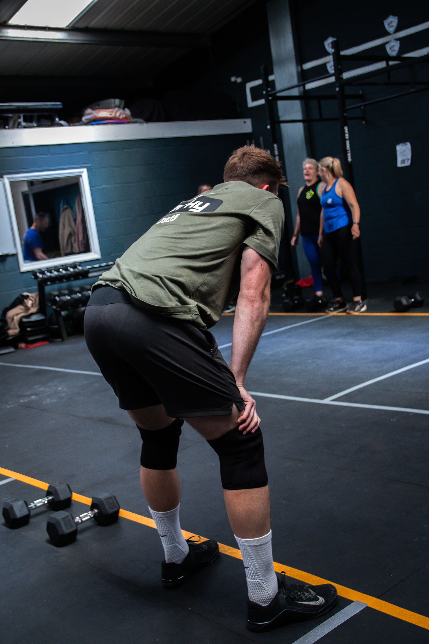 Athlete in knee sleeves and olive tank top stretches before workout with dumbbells visible on gym floor.