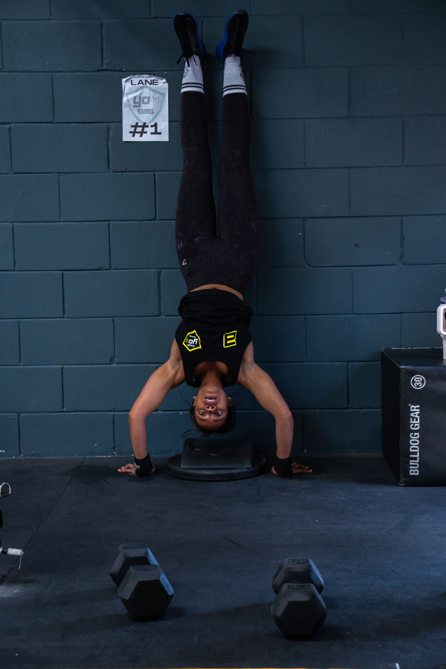 A person performs a handstand against a blue concrete wall with dumbbells on the floor in a gym setting.
