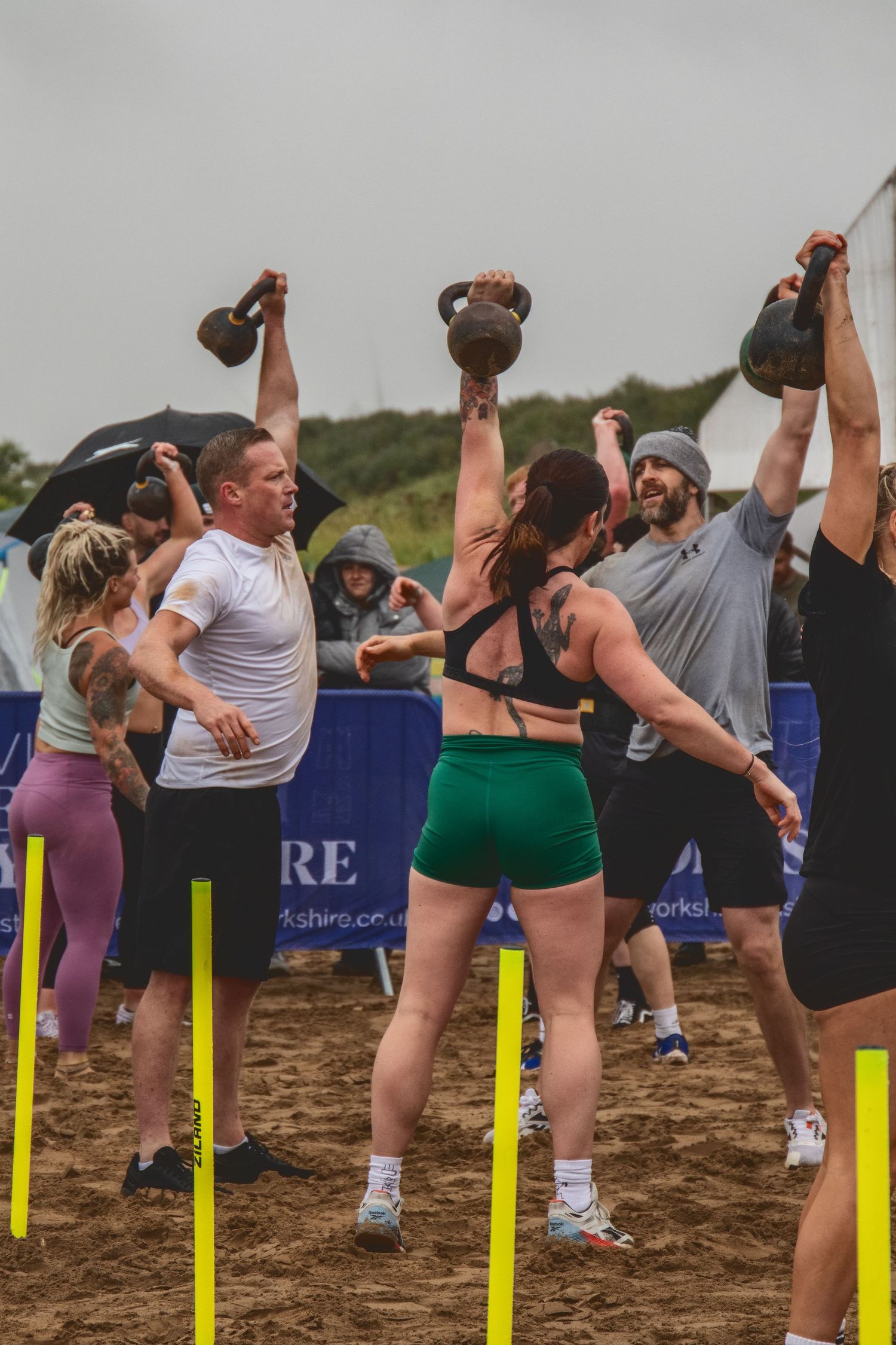 A group of athletes perform kettlebell exercises during an outdoor fitness class on sandy terrain.