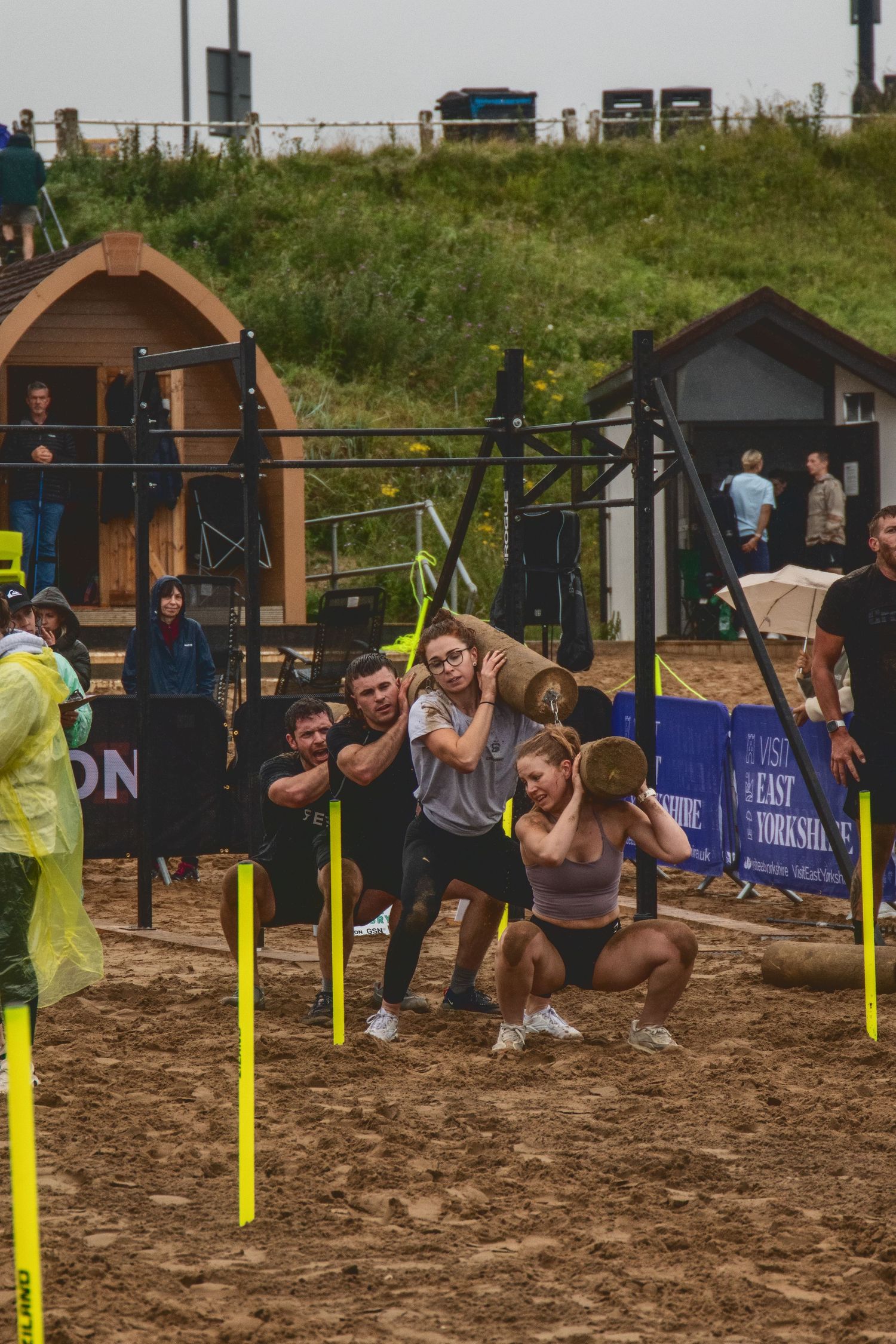 Athletes and spectators gather at an outdoor fitness competition on sandy ground with training equipment and obstacles.