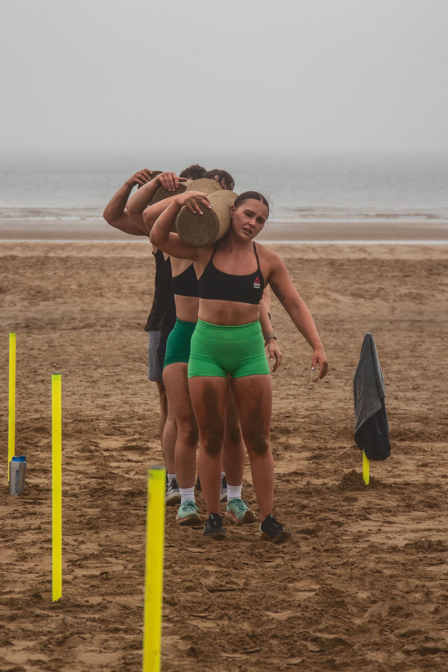 Athletes in green workout gear train together on a foggy beach with yellow posts marking their exercise area.
