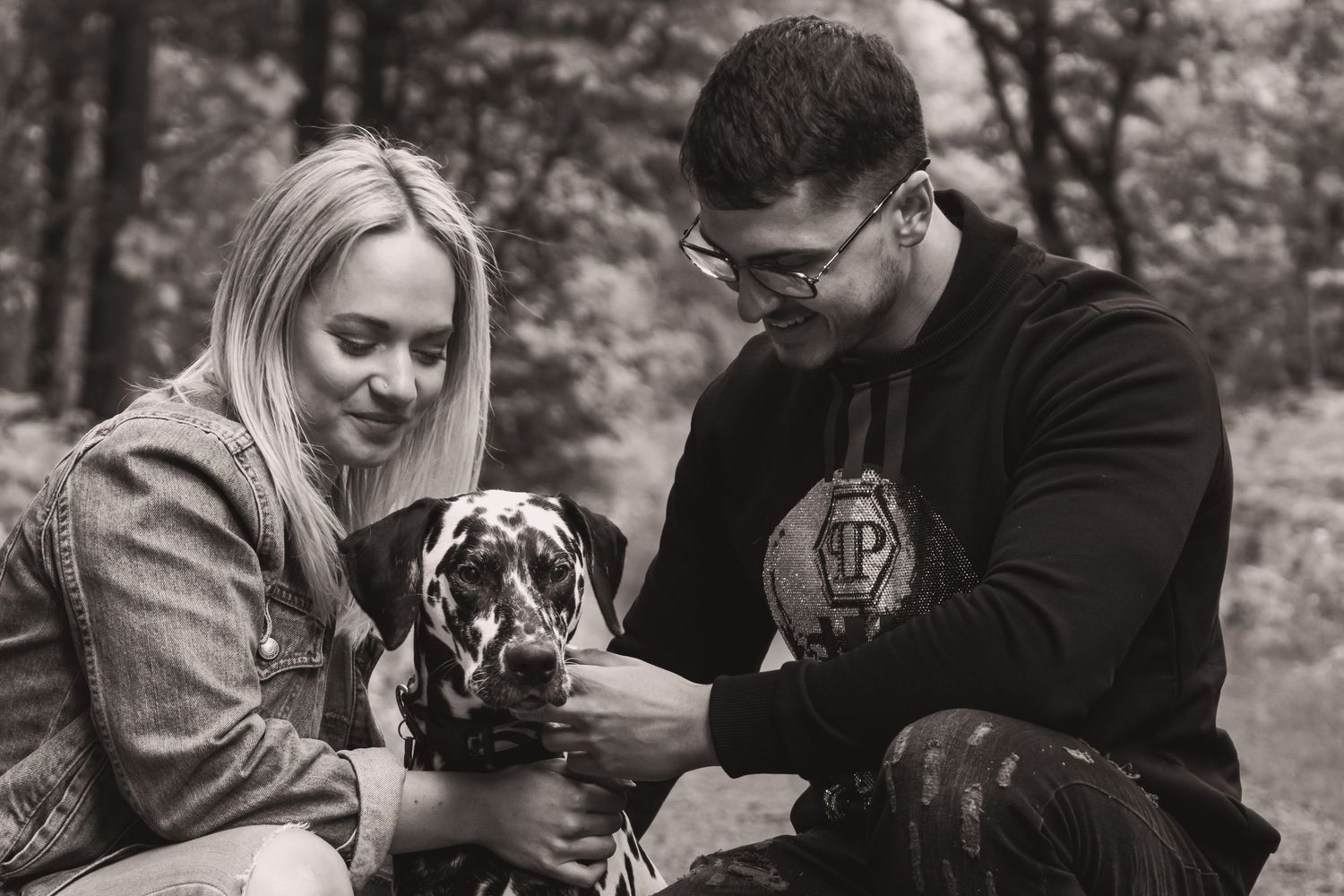 A couple sits outdoors with their spotted Dalmatian dog in a black and white photo.