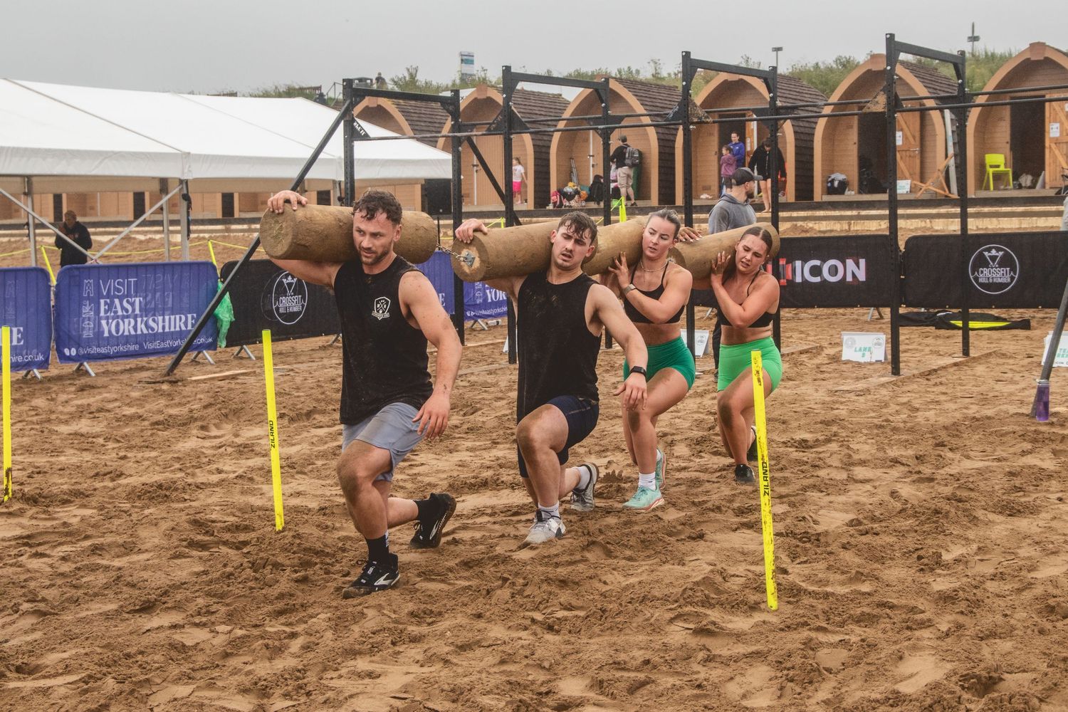 Athletes compete in an outdoor obstacle course race crawling through mud beneath low ropes and barriers.