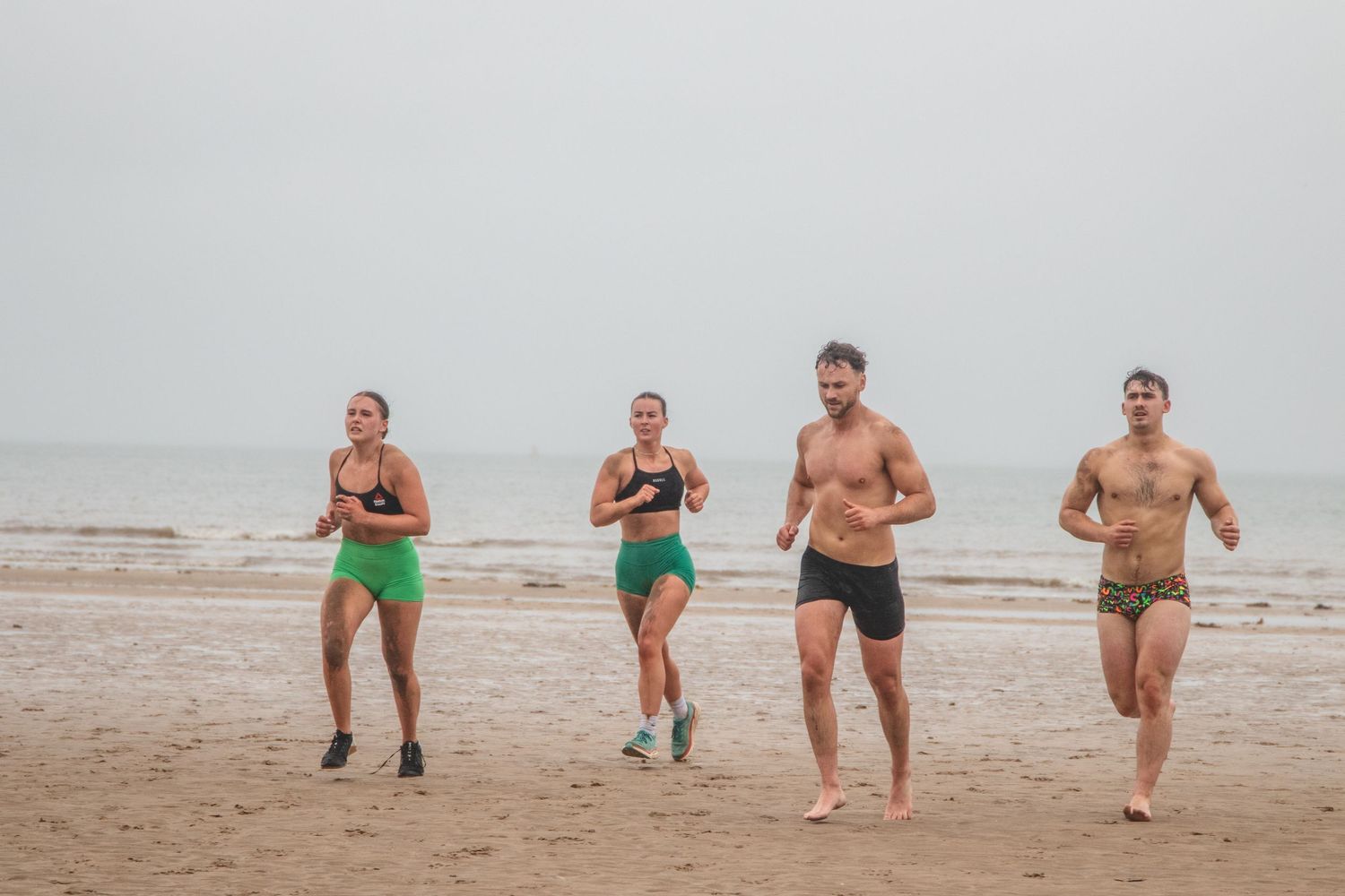 A group of adults perform a bare-chest dance on the beach while wearing green shirts and swimsuits.