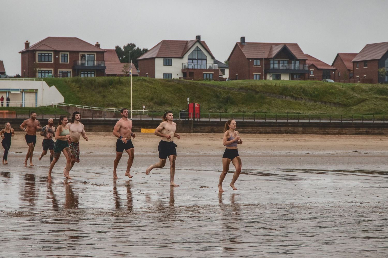 A group of people running barefoot in shallow beach water with coastal homes visible on a hill in the background.