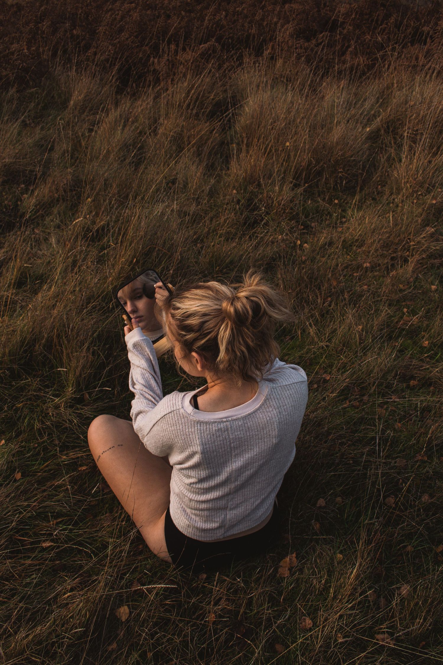 Person in a cream knit sweater sits thoughtfully in tall autumn grass during golden hour lighting.