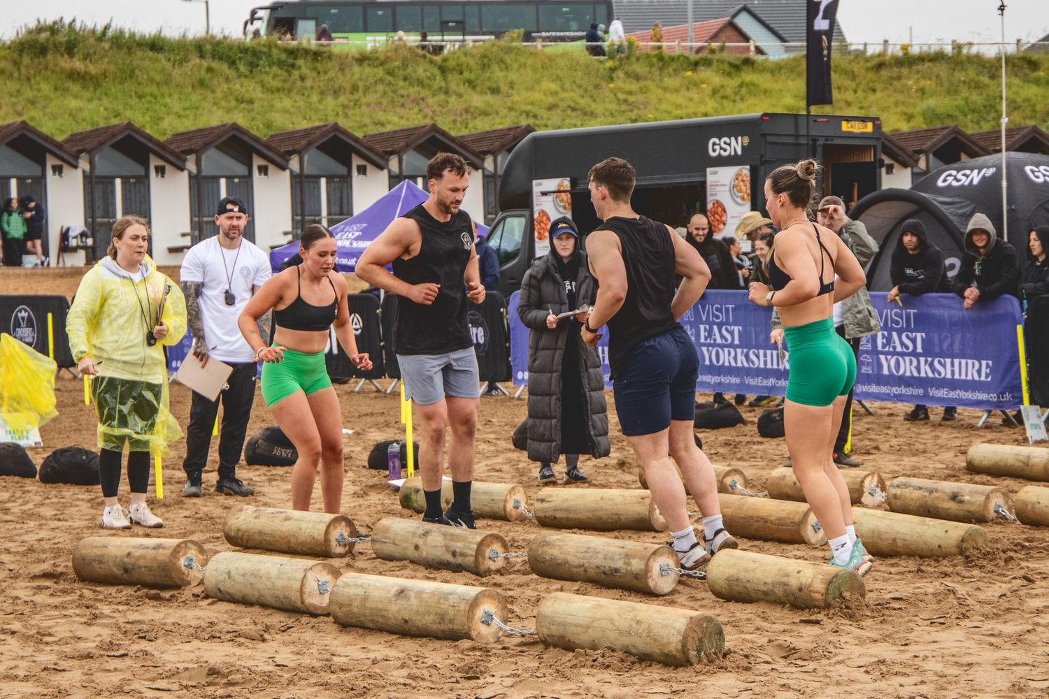 Participants roll and carry large wooden logs across a dirt field during a competitive outdoor strength event.