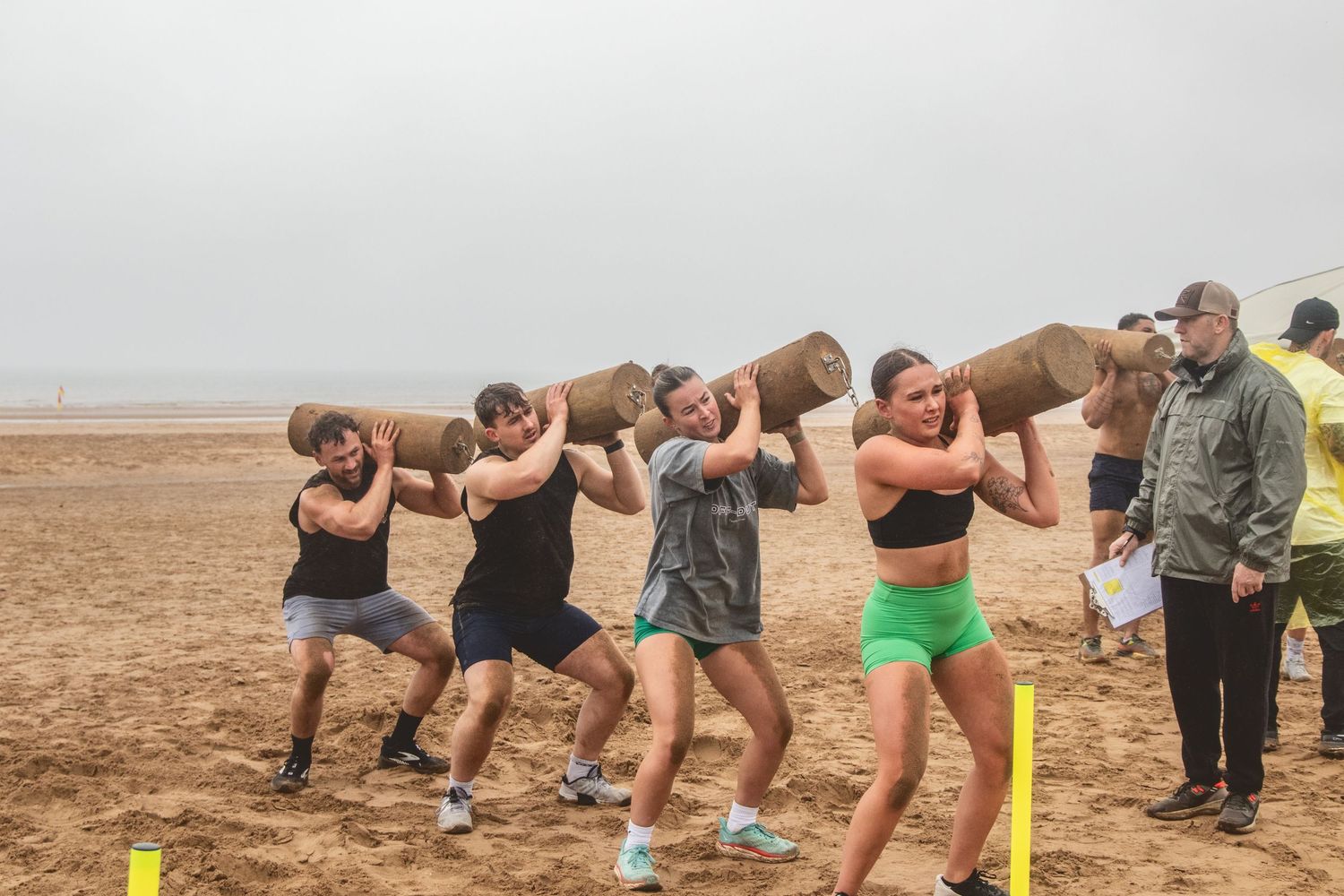 People performing synchronized squats with wooden logs on their shoulders at a sandy beach during group workout.