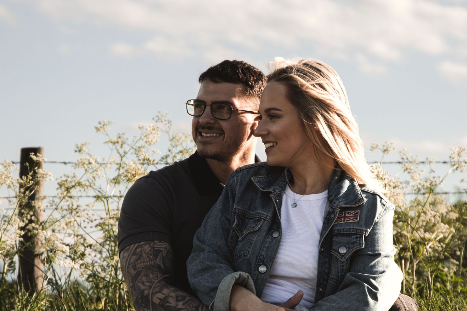 Couple embraces outdoors on a sunny day with tall grass and blue sky in background.