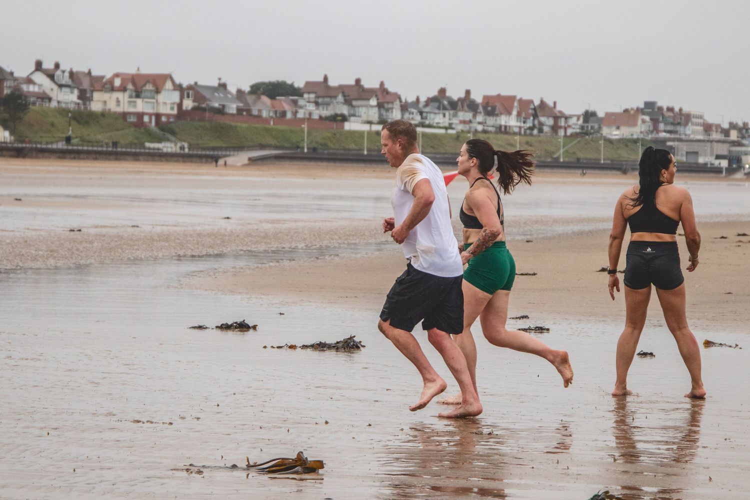 Three people jogging together along a sandy beach with coastal houses visible in the background.