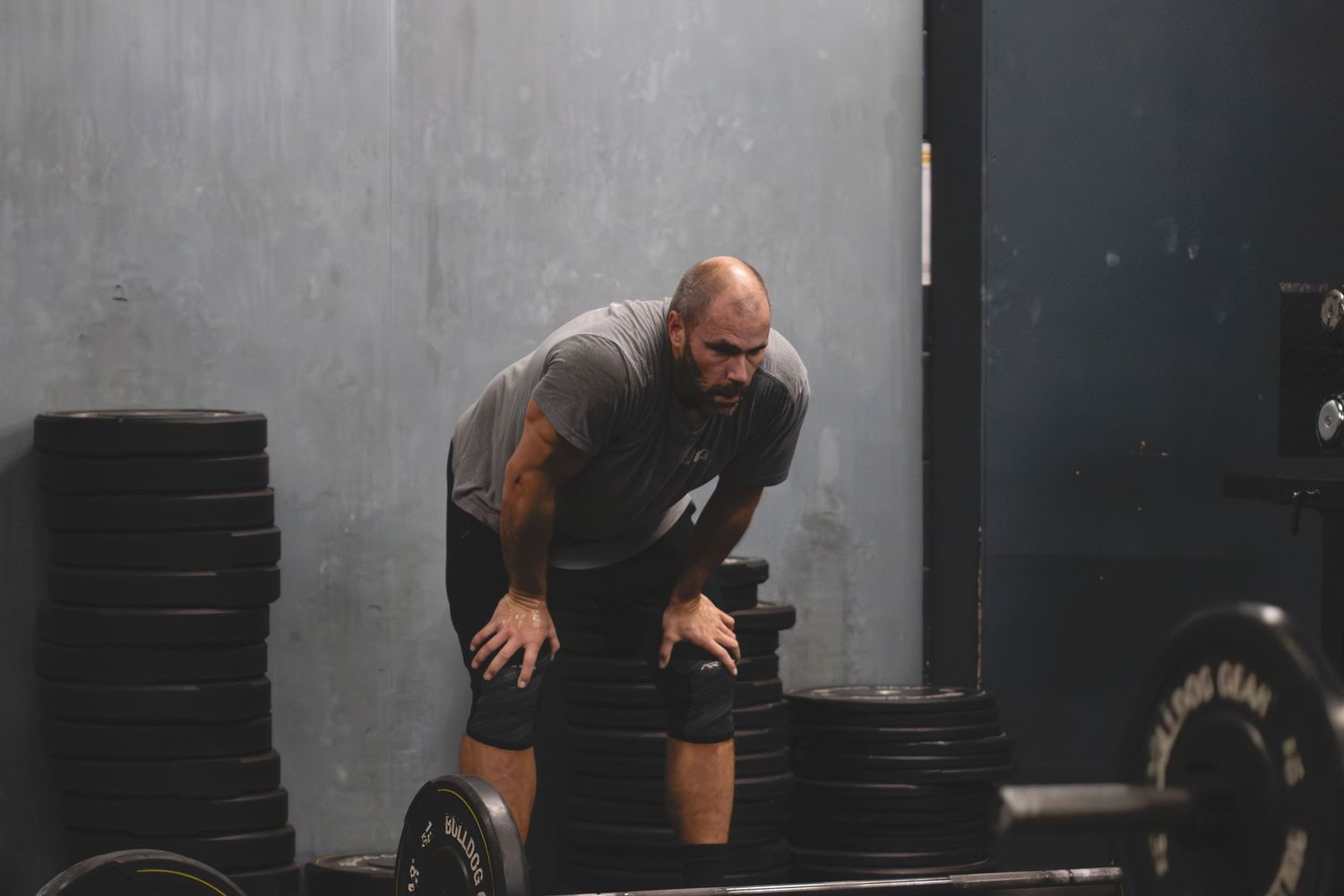 Tired athlete catching his breath next to a barbell during an intense CrossFit workout in a dark gym.