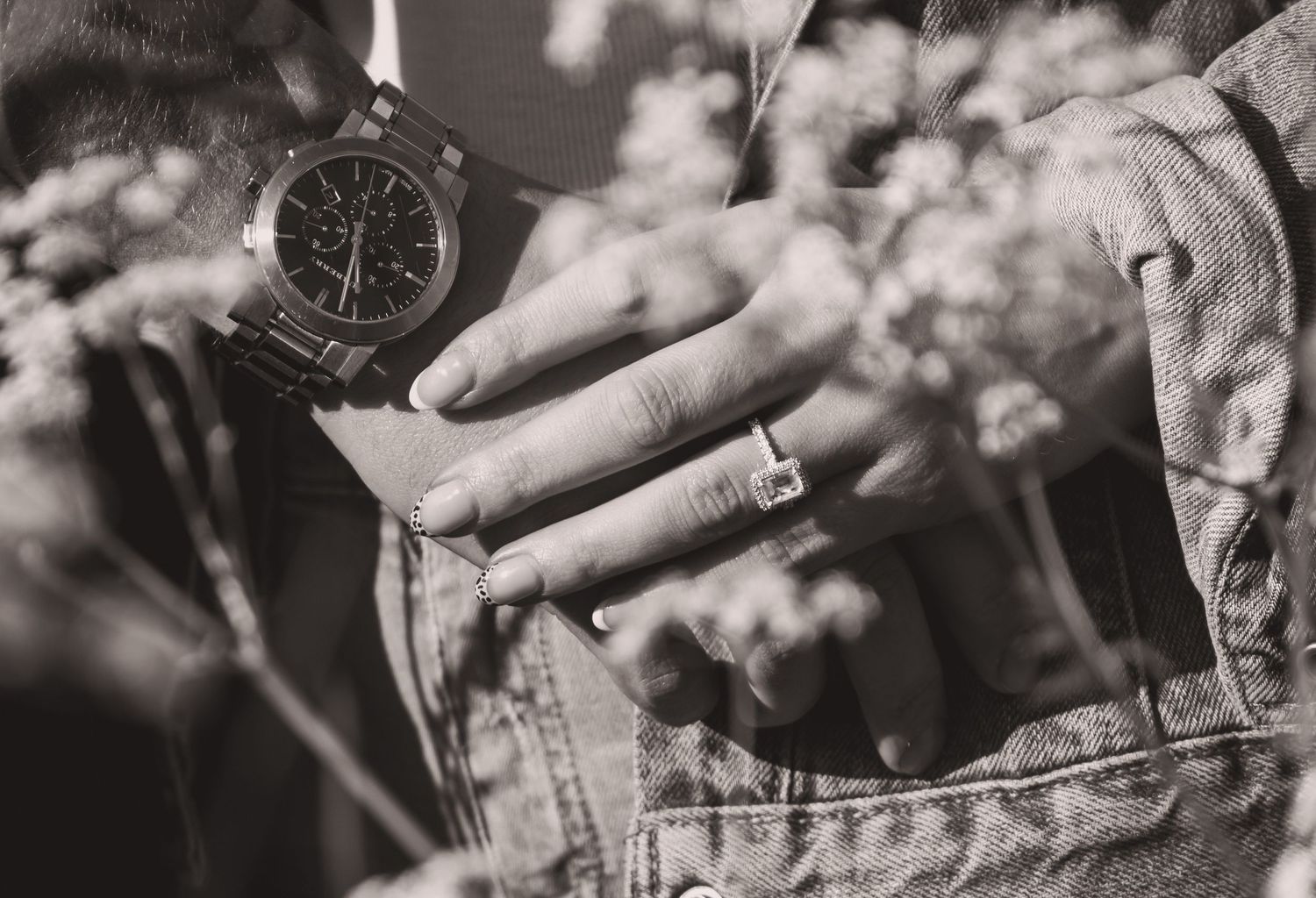 Monochrome close-up of hands showing watch and ring details against floral background.