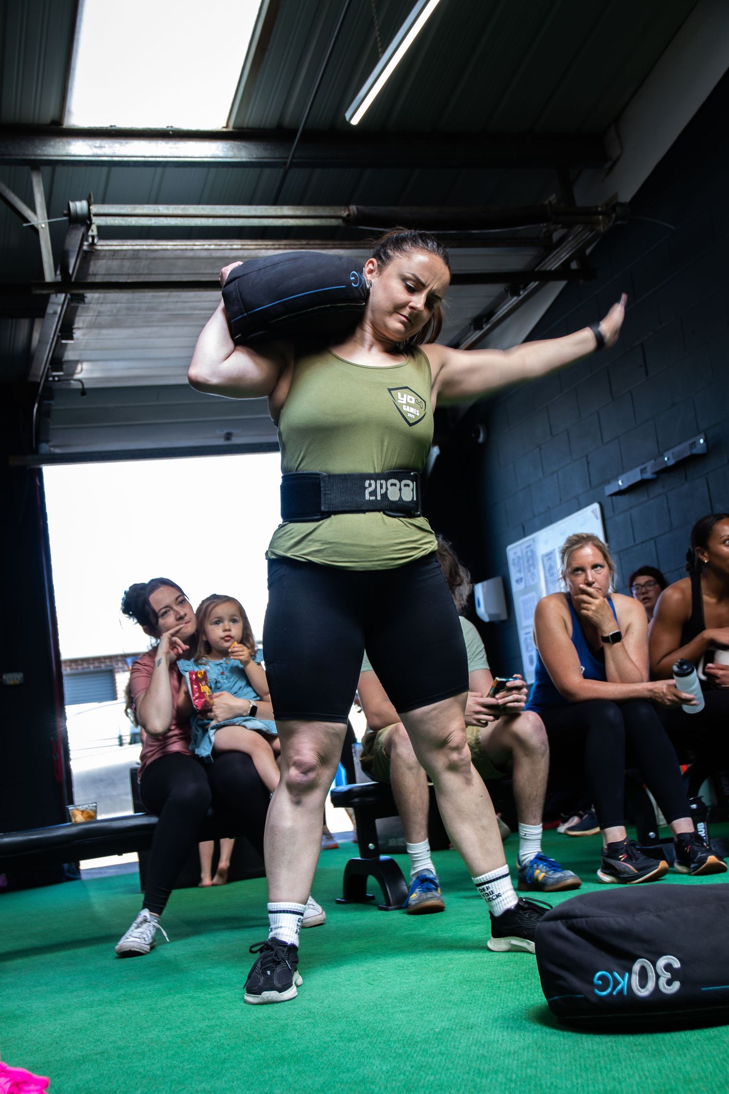An athlete performs a sandbag exercise on green turf flooring during a CrossFit-style workout class.