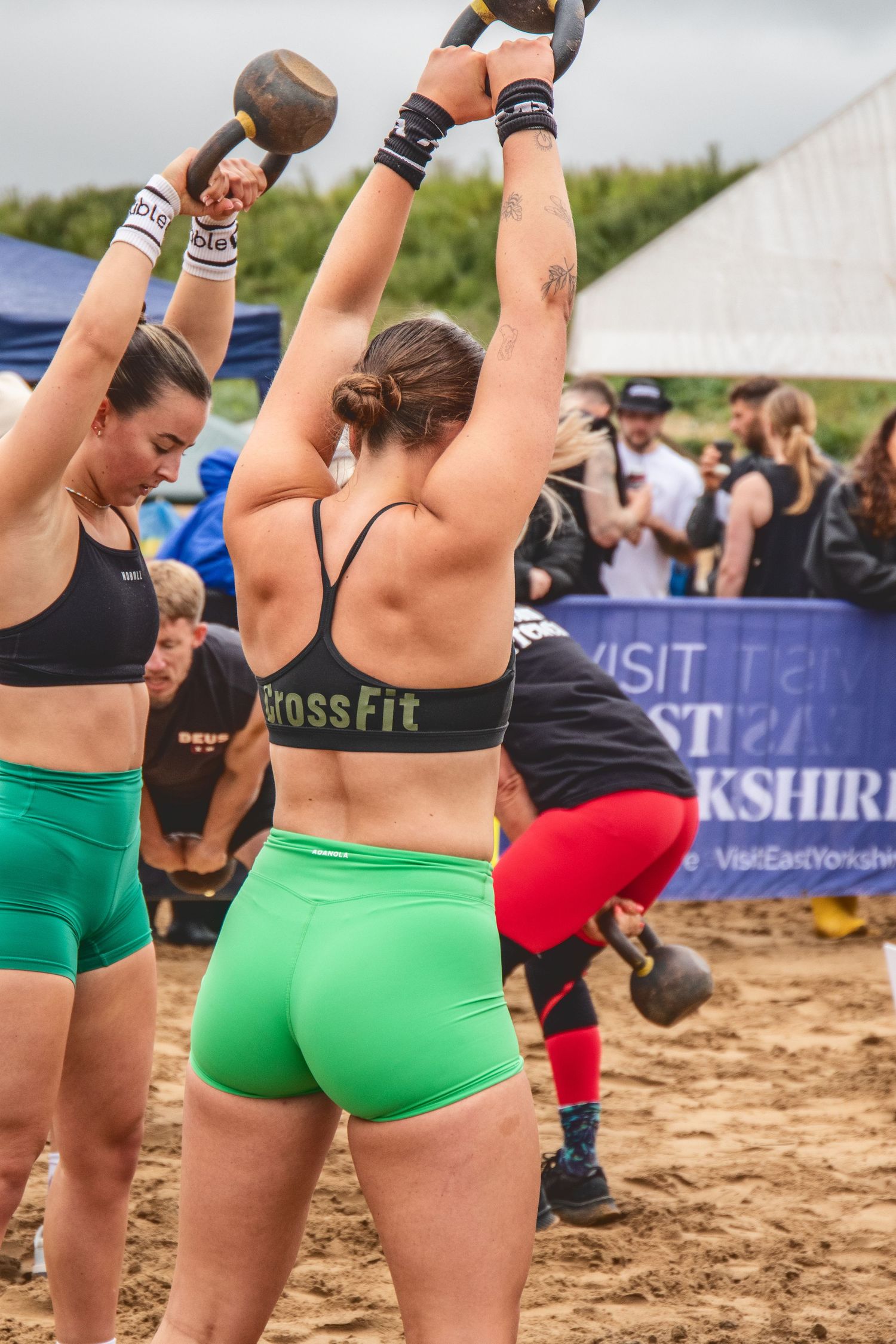 Athletes in green shorts and sports bras compete at a CrossFit competition on a sandy beach area.