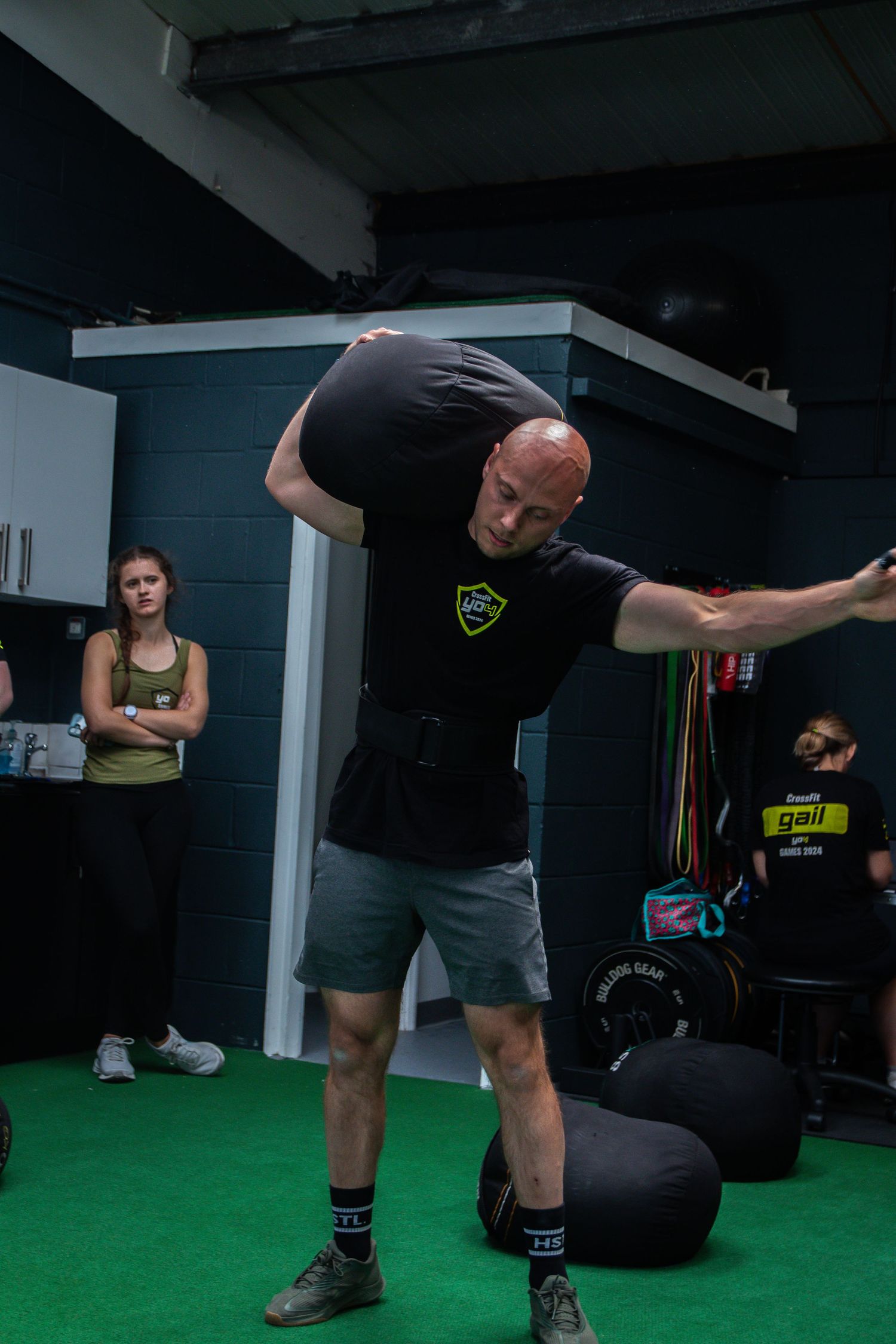 Athlete performs sandbag training exercise on green turf floor in gym setting with training equipment visible.