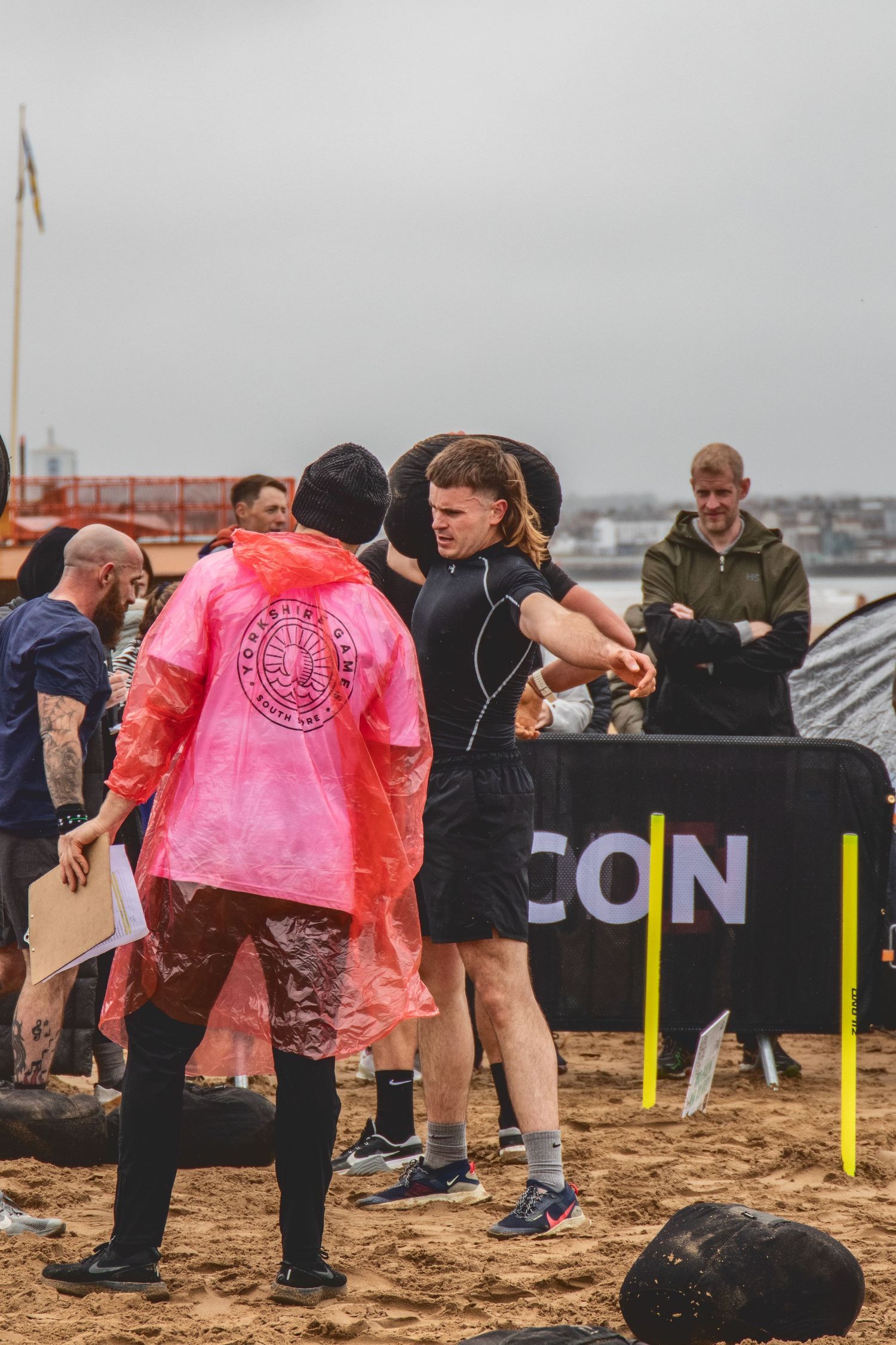 Athletes compete in an outdoor obstacle course on sandy terrain with spectators watching from behind barriers.