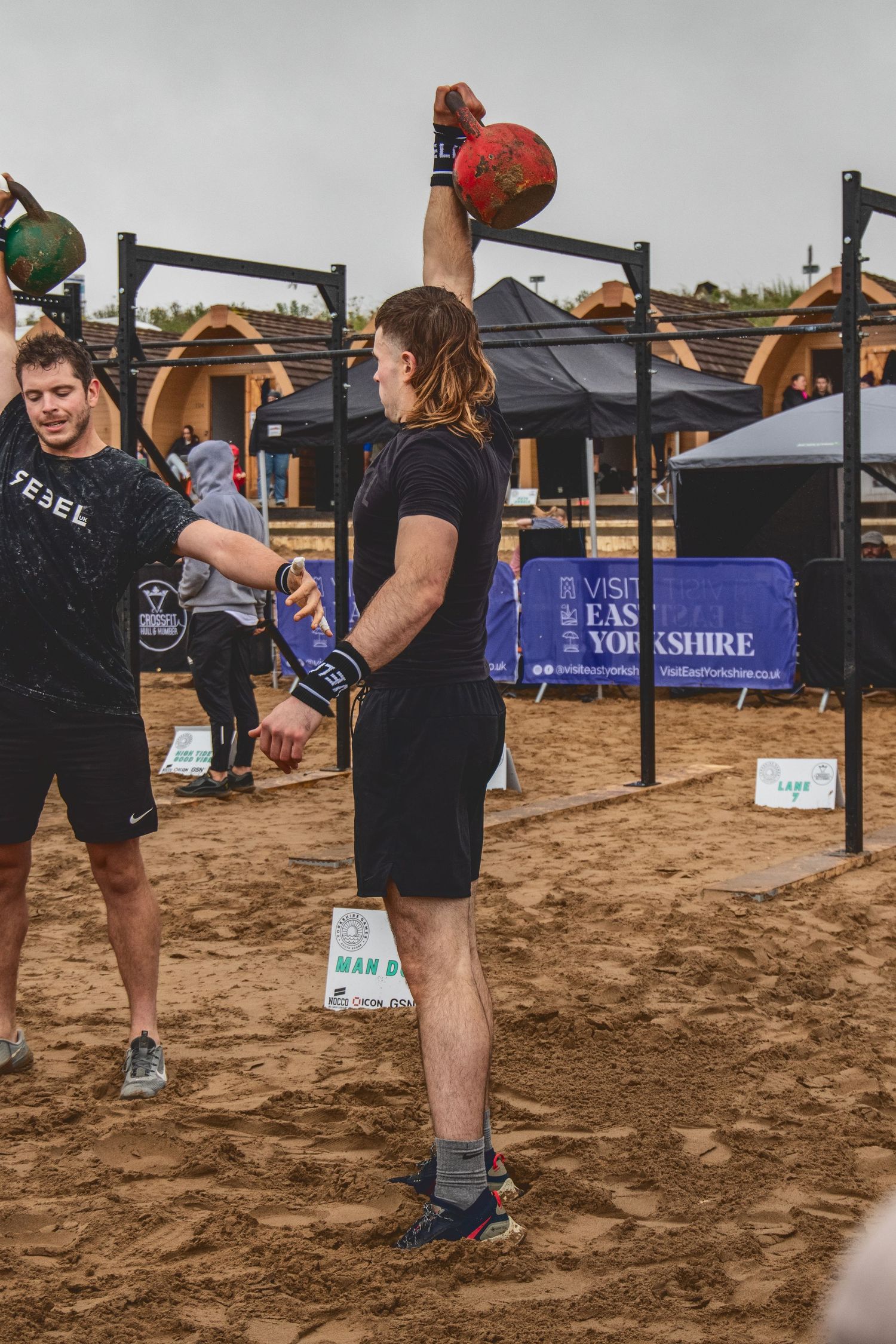 Athletes perform burpee box jump exercises on sandy terrain during an outdoor fitness competition.