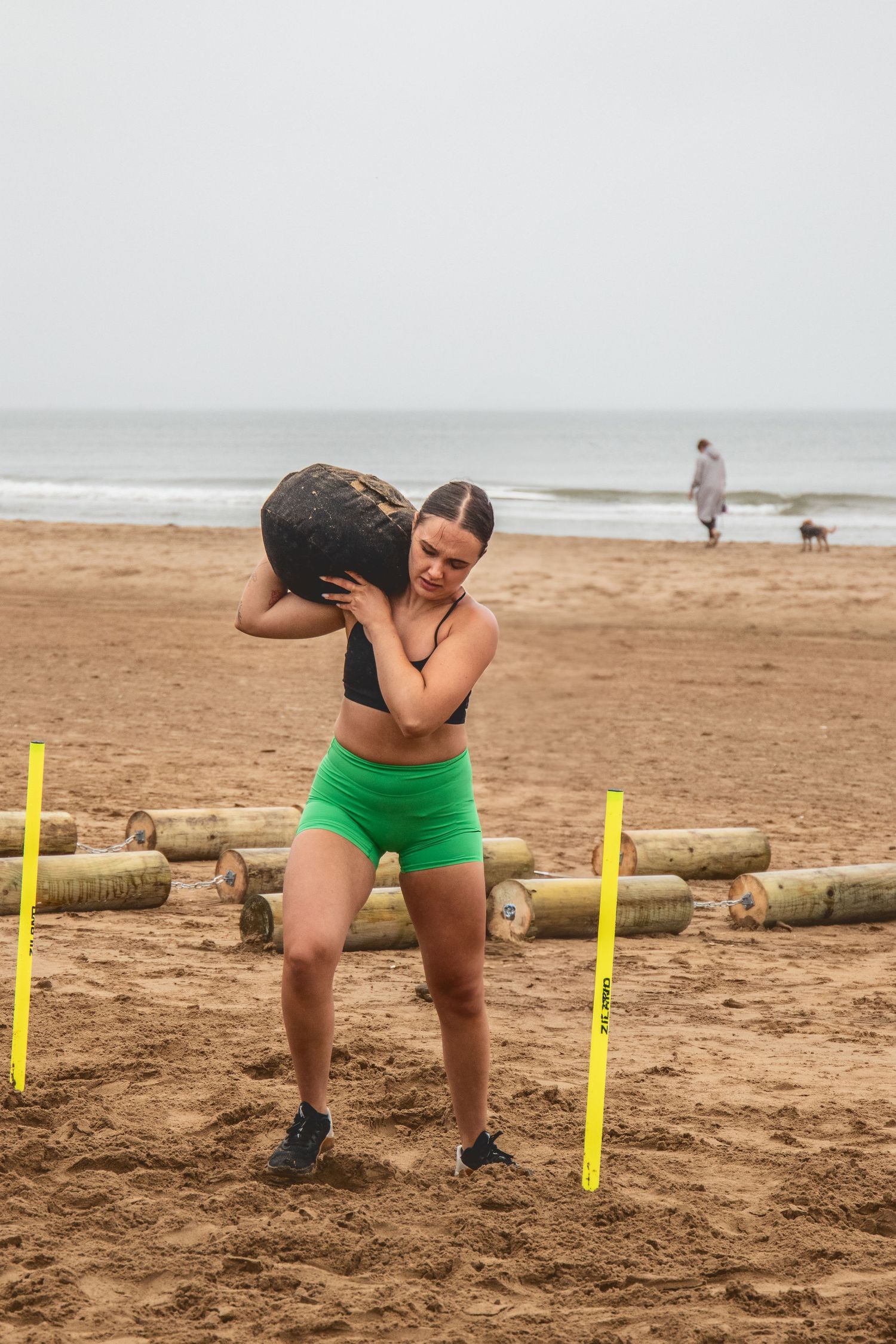 Athlete in green shorts performing sandbag exercises during beach workout with yellow markers on sandy shore.