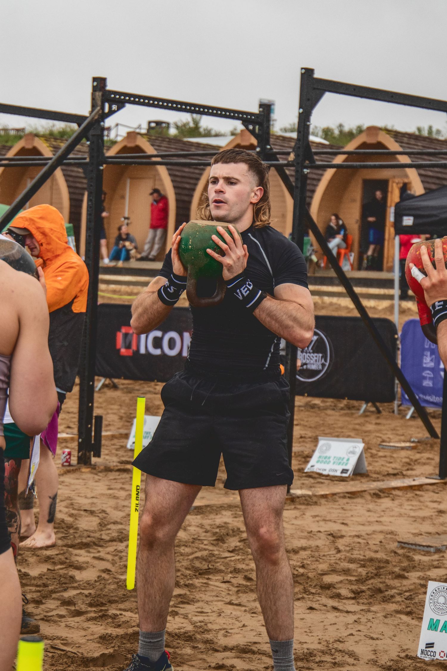 Athlete in black workout gear performs medicine ball exercise at outdoor CrossFit competition on sandy ground.