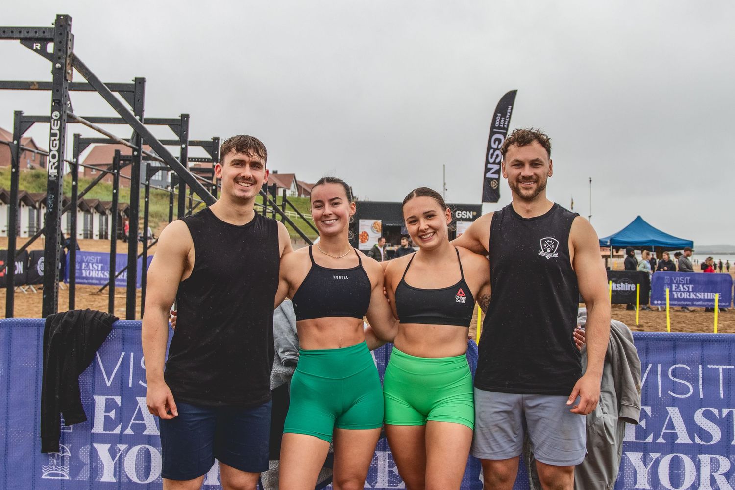 Athletes in workout attire stand together at an outdoor fitness event with exercise equipment visible in the background.