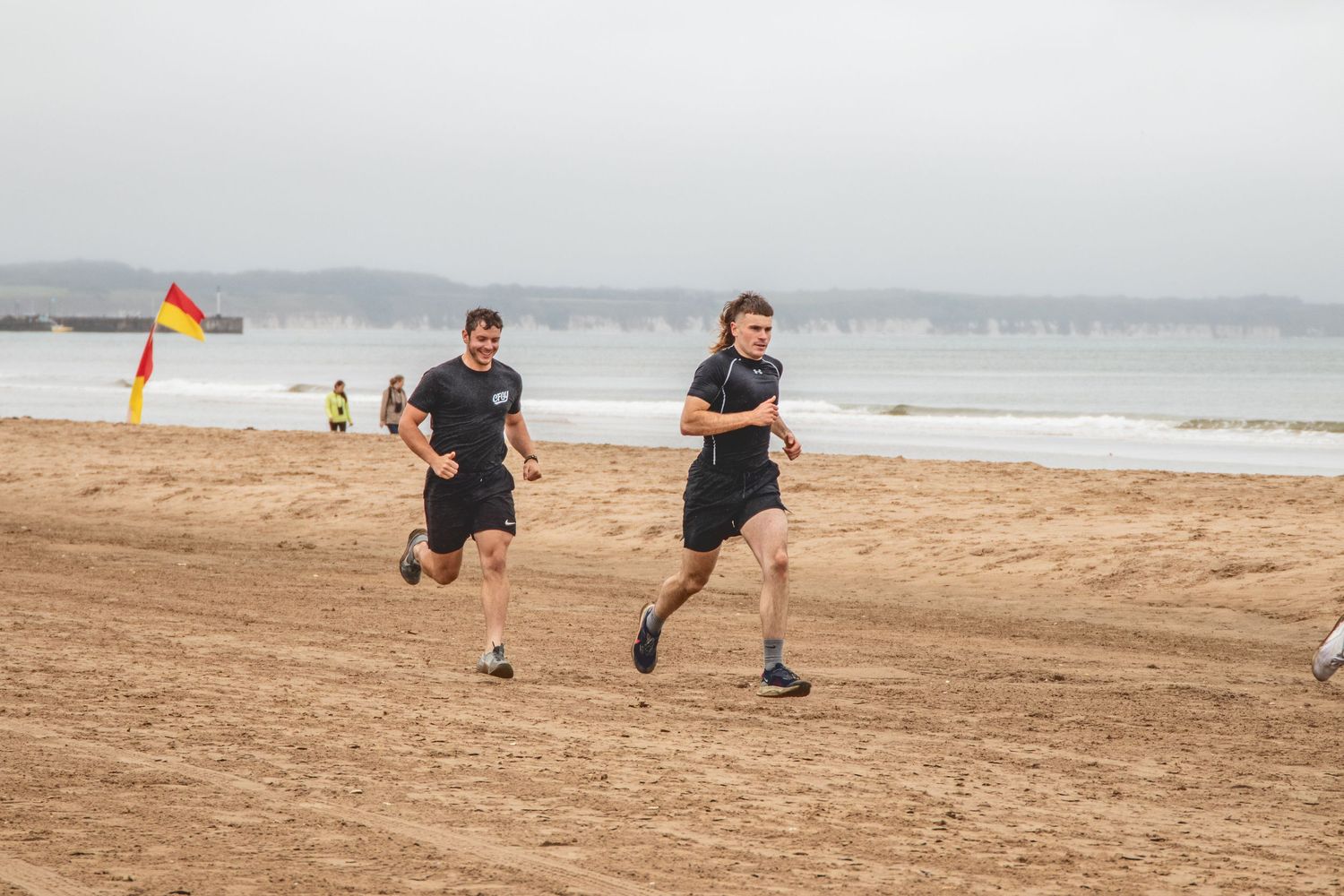 Two runners in black athletic wear train together on a sandy beach with ocean and hills visible in the background.