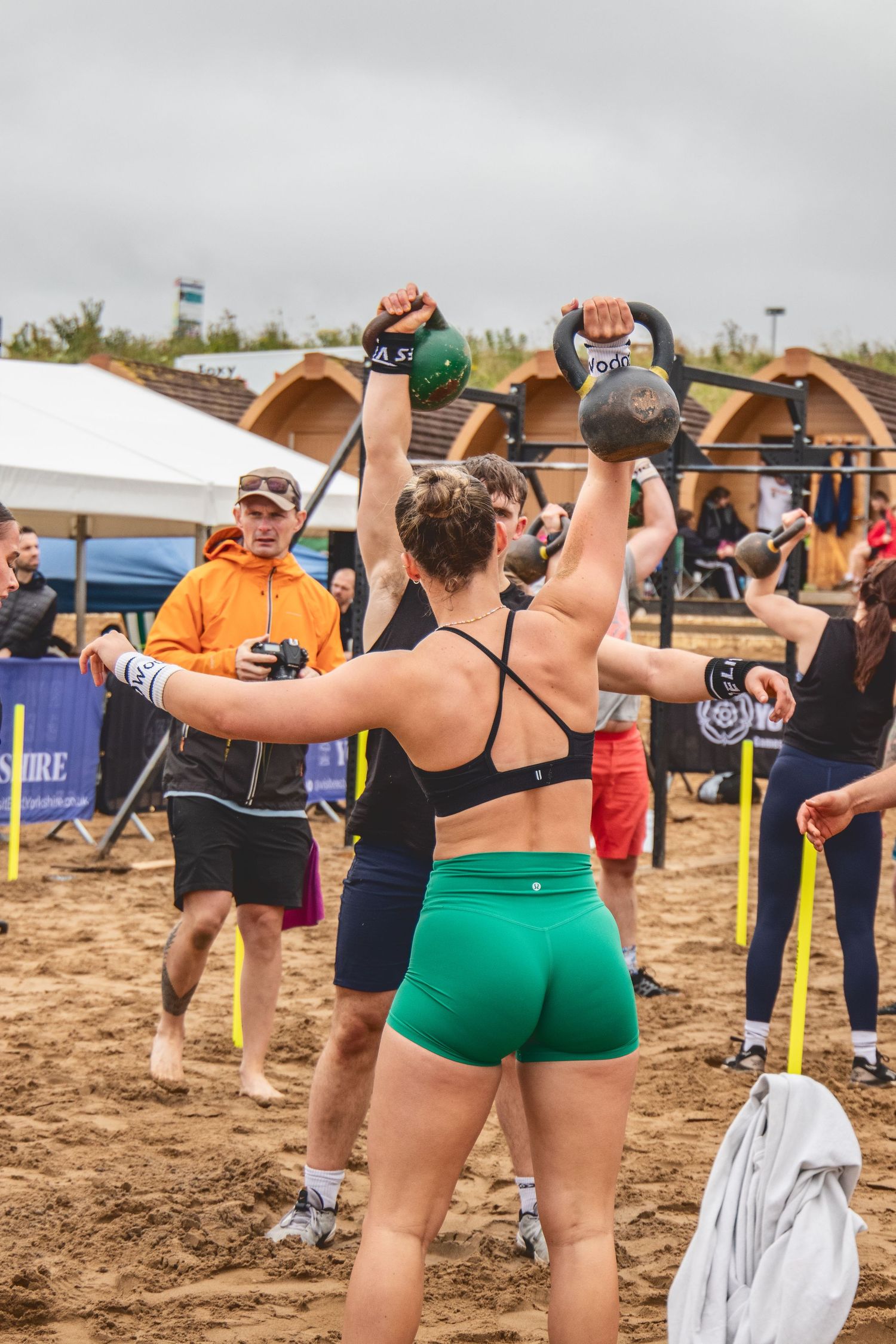 Athletes perform kettlebell workouts during an outdoor fitness competition on sandy terrain with spectators watching.