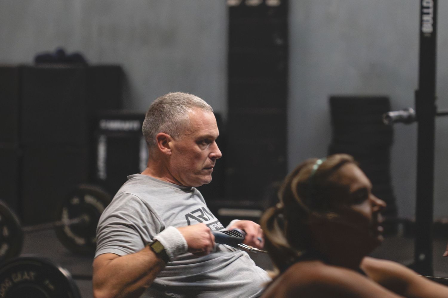 Person in gray t-shirt working out with dumbbells in dark gym setting with exercise equipment in background.