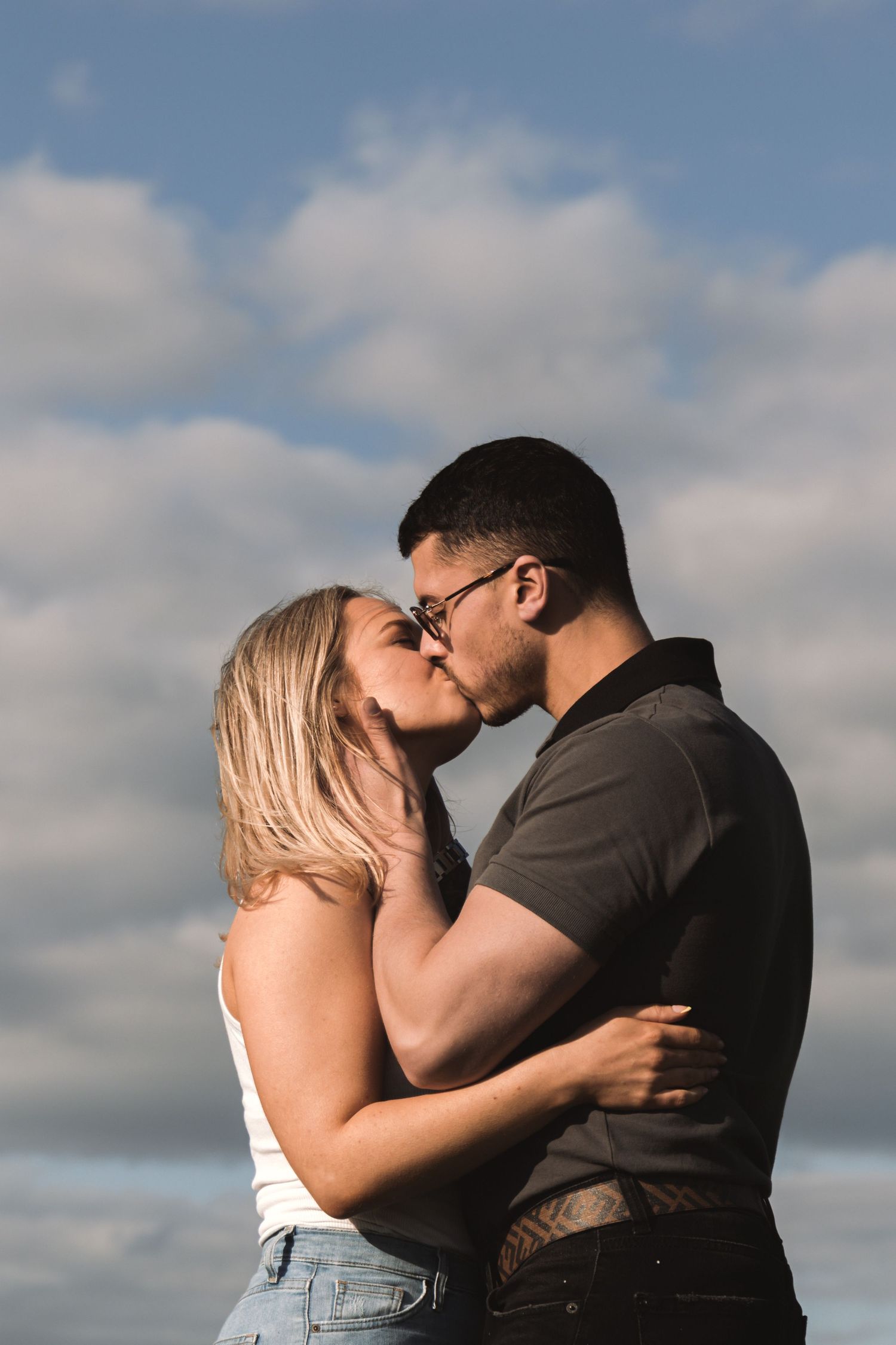 Romantic couple embraces and kisses in front of a cloudy sky during an outdoor photoshoot.
