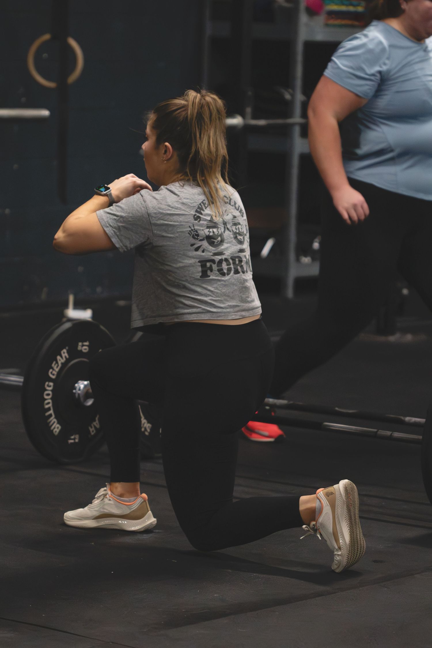 Someone in workout gear performs a lunge exercise next to a barbell in a dark gym setting.
