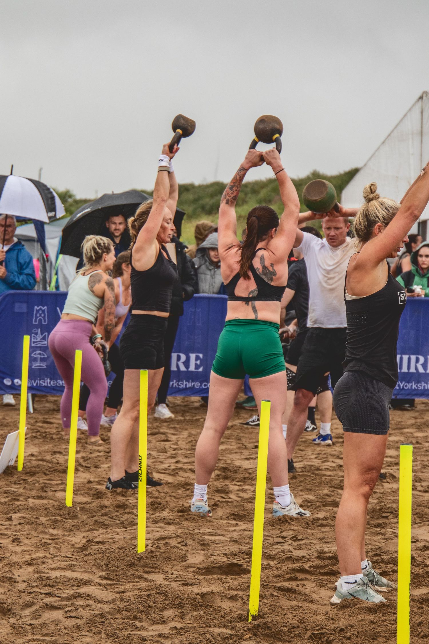 Athletes lifting kettlebells during an outdoor fitness competition on sandy beach terrain with yellow markers.