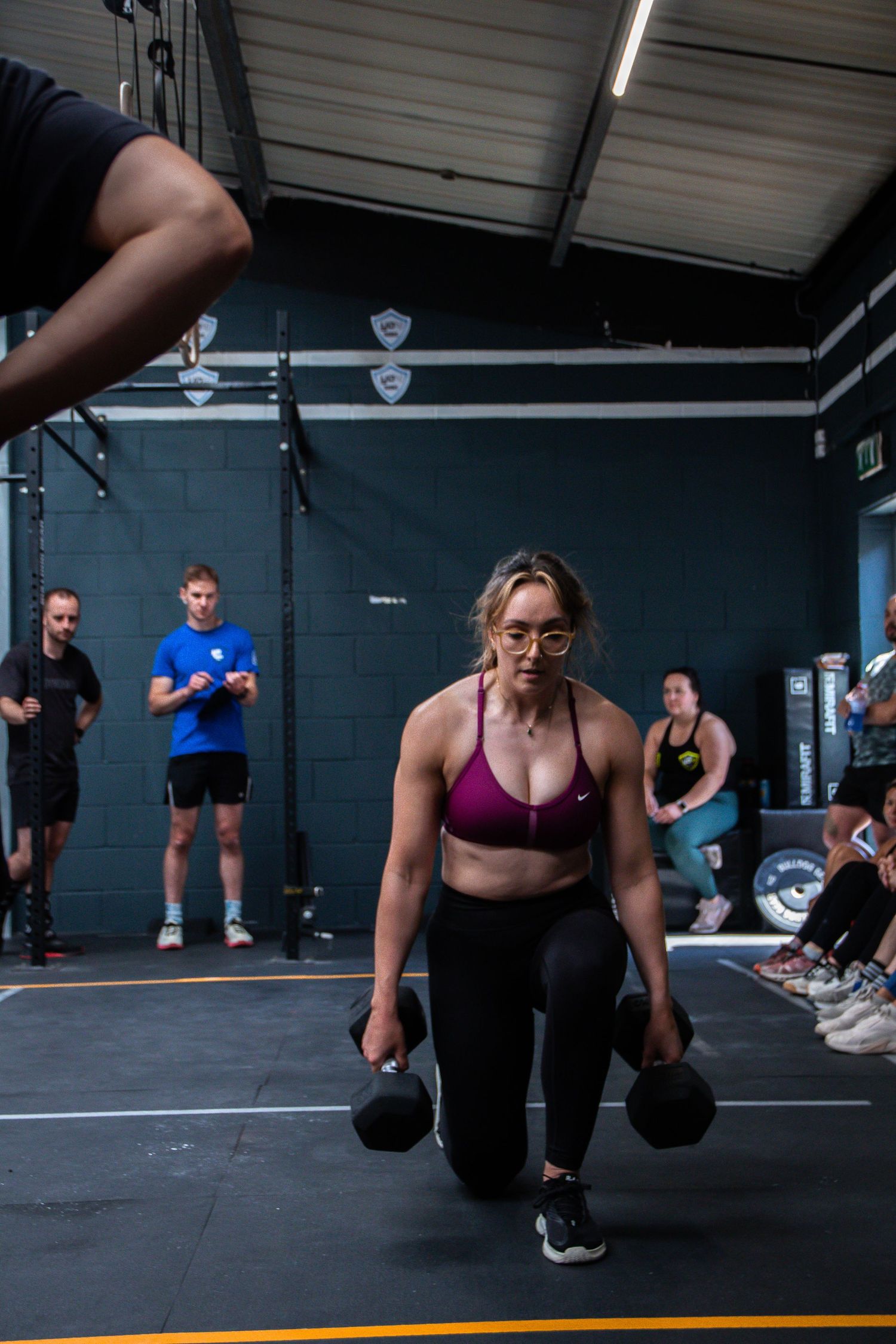A sequence of CrossFit workout movements showing athletes performing dumbbell exercises on a dark gym floor.