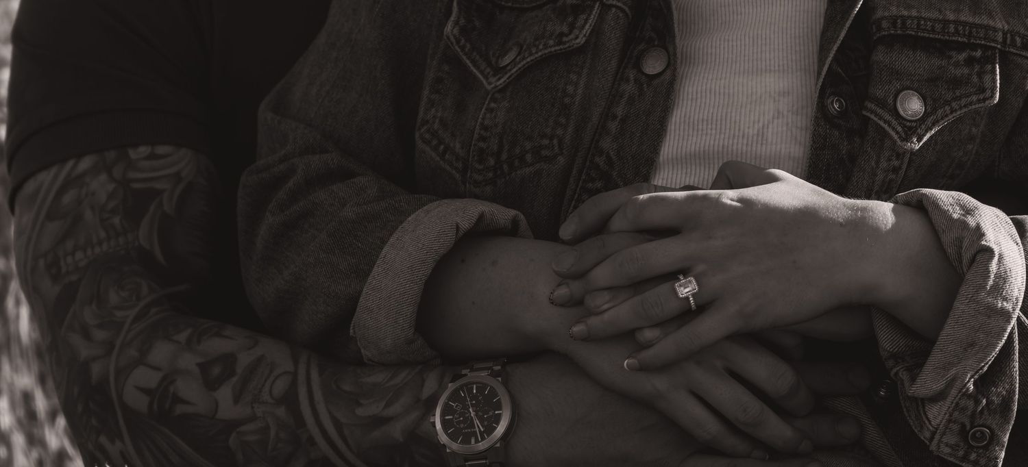 Close-up black and white photo of hands with engagement ring and tattooed arm.