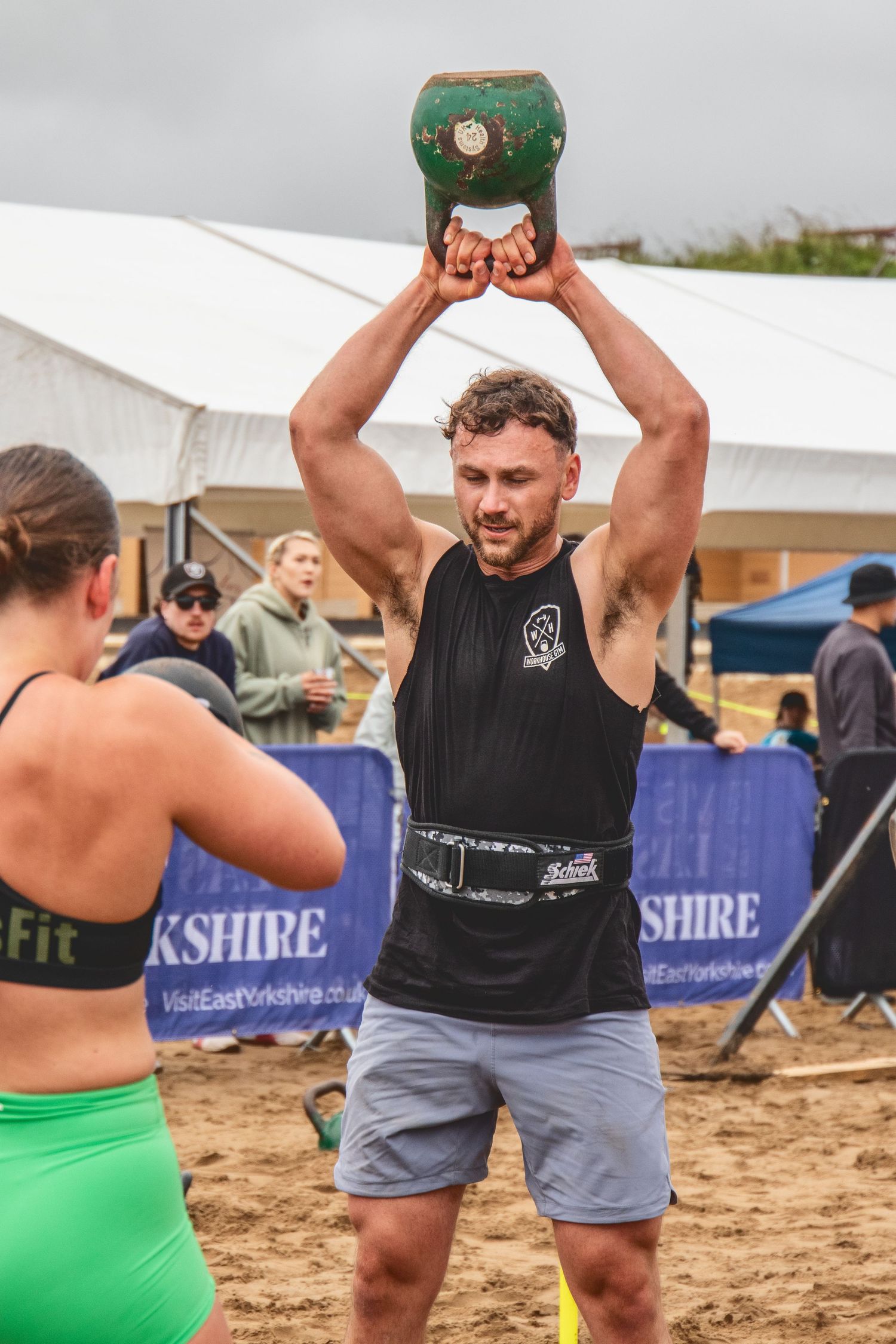 Athlete lifting heavy kettlebell overhead during outdoor CrossFit competition on sandy ground with event tent in background.