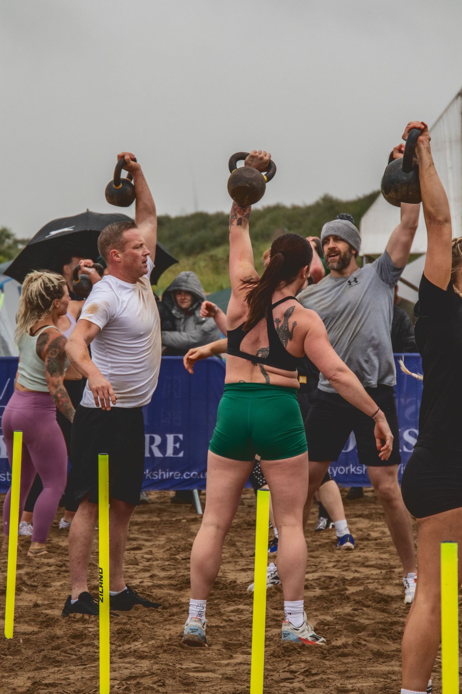 Athletes lifting kettlebells during an outdoor fitness competition on sandy ground with yellow markers.