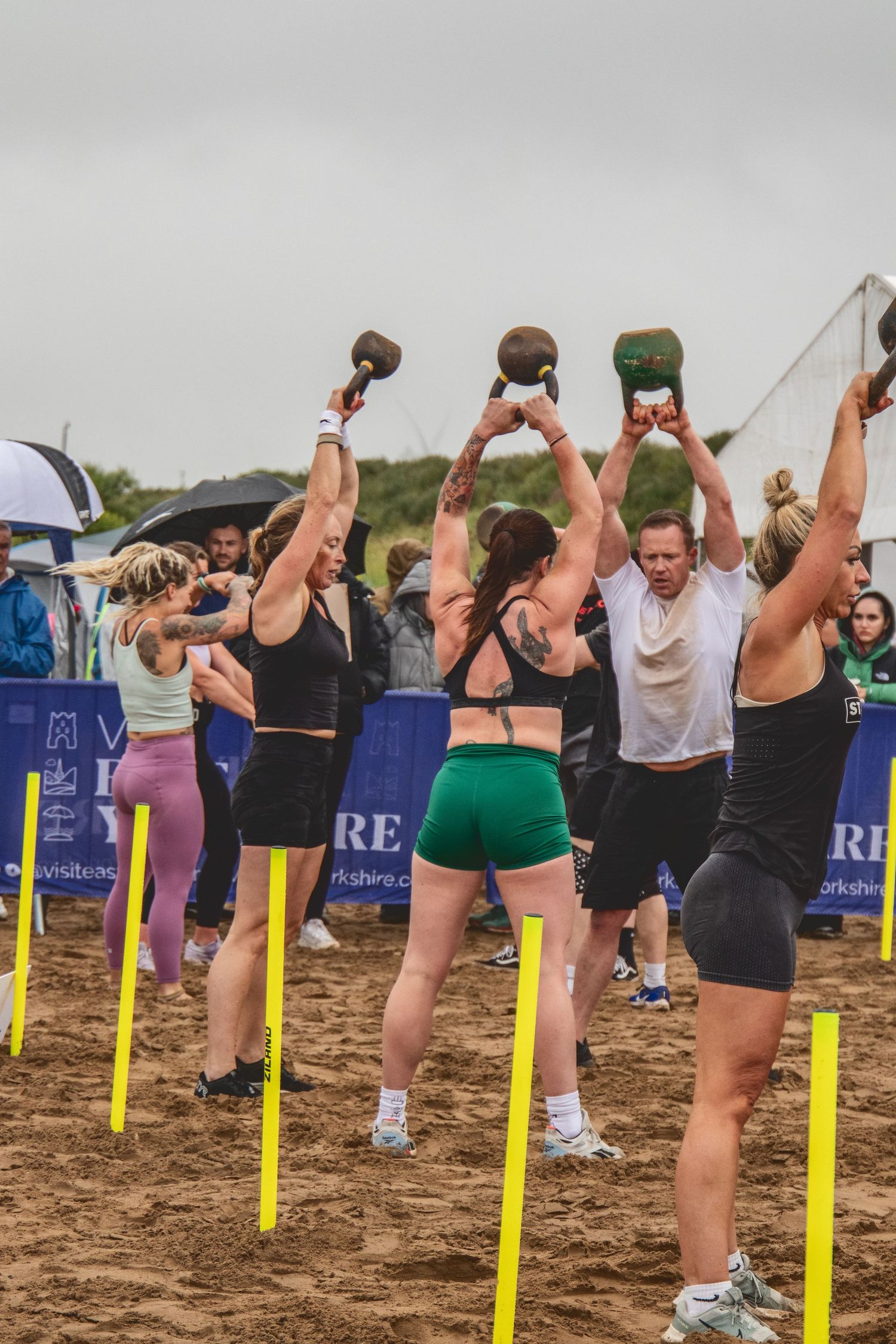 A group of people perform a synchronized overhead weight lifting exercise with medicine balls on a beach.