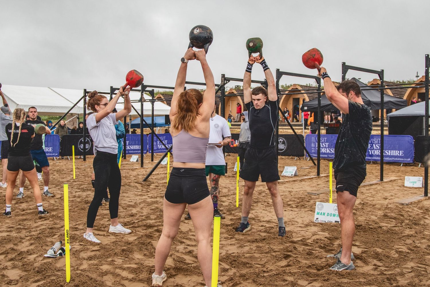 Athletes perform overhead lifts with medicine balls during an outdoor fitness competition on sandy terrain.